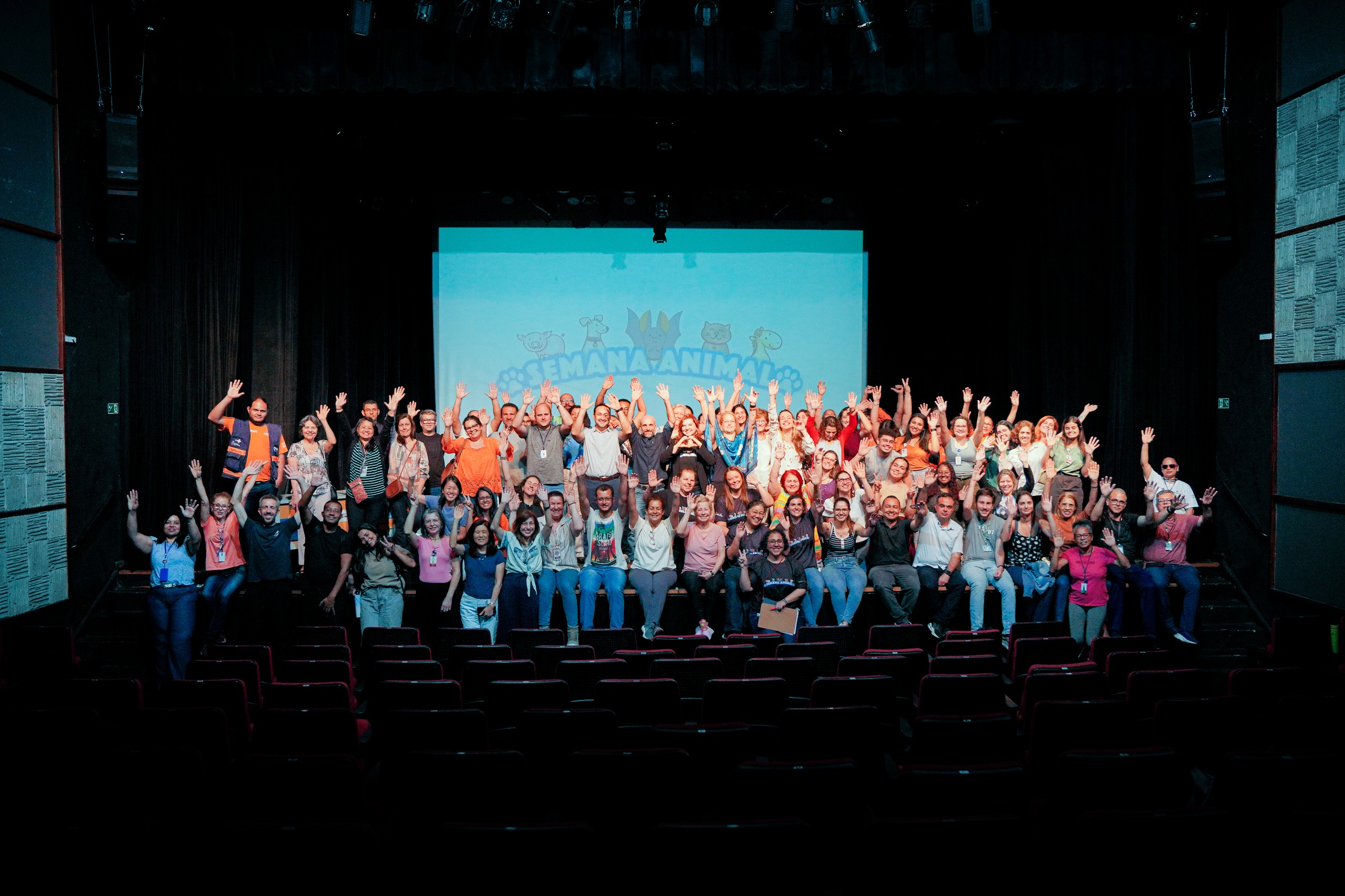 A foto mostra um grande grupo de pessoas reunidas em um teatro. Elas estão posicionadas no palco, em duas fileiras, e muitas levantam os braços em gesto de celebração e alegria. O auditório está escuro, com as poltronas vazias visíveis em primeiro plano.  No fundo, há uma tela de projeção com ilustrações de animais sorridentes e a frase “Semana Animal” em letras azuis, reforçando o clima festivo e o tema do encontro.  As pessoas no palco são diversas em idade, gênero e aparência, vestindo roupas casuais e coloridas. O ambiente transmite união, engajamento e espírito de confraternização, como se fosse o encerramento de um evento coletivo.