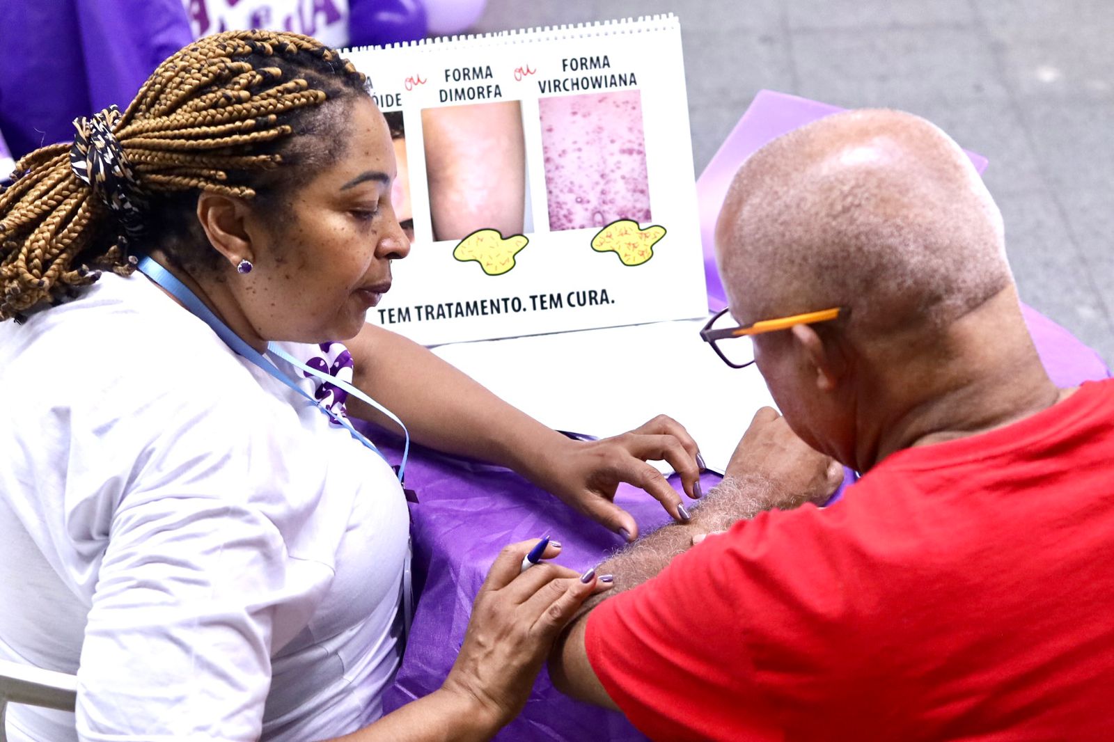 A imagem mostra uma mulher negra, com cabelos trançados, usando camiseta branca realizando uma avaliação na pele de um homem, que veste uma camiseta vermelha e óculos com armação laranja. Ao fundo, há um cartaz com imagens de diferentes formas de hanseníase, intituladas