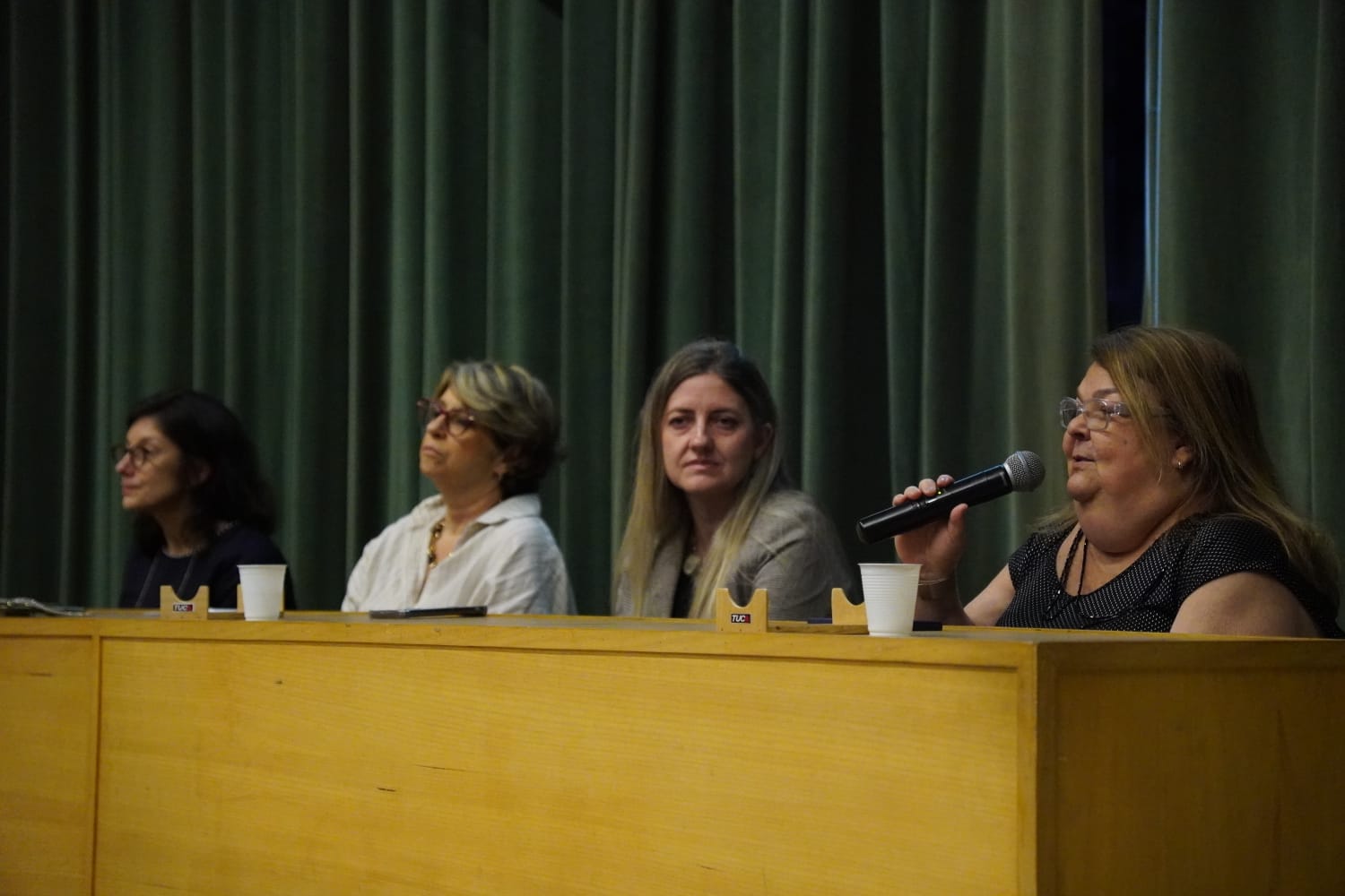 A imagem mostra uma mesa de debate com quatro mulheres sentadas atrás de uma bancada de madeira clara. Ao fundo, há uma cortina verde escura que ocupa. Da esquerda para a direita: A primeira mulher tem cabelo escuro, usa óculos e veste uma blusa preta. A segunda mulher tem cabelo curto e claro, usa óculos e veste uma camisa branca. A terceira mulher tem cabelos longos e loiros, veste um blazer claro. A quarta mulher tem cabelos castanhos claros, usa óculos e veste uma blusa preta, e está falando no microfone. 