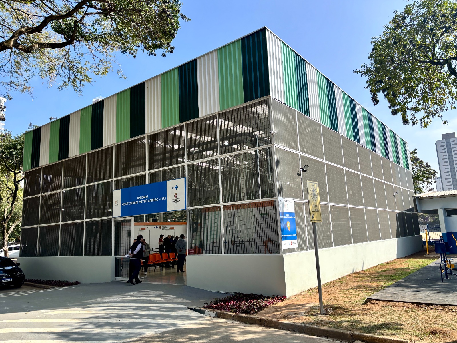 A imagem mostra a fachada de um prédio público moderno, identificado como Unidade Monte Serrat Metrô Carrão – CIES, pertencente à Secretaria Municipal da Saúde de São Paulo.  A construção é retangular, com paredes revestidas por placas metálicas verticais em diferentes tons de verde e branco na parte superior, que dão um aspecto visual dinâmico e colorido. A parte inferior é cercada por grades metálicas, permitindo ventilação e visibilidade interna. Na entrada principal, há uma placa azul com o nome da unidade e o logotipo do SUS, além do brasão da Prefeitura de São Paulo.  Algumas pessoas estão entrando no prédio, onde se pode ver cadeiras de espera de cor laranja. Do lado de fora, o piso é de concreto claro, com marcações de acessibilidade e uma faixa de pedestres pintada. À direita, há um pequeno espaço com solo de terra batida e grama, além de equipamentos que lembram aparelhos de ginástica ao ar livre. Também há um poste com uma placa amarela indicando “Área Escolar”.  Ao fundo, aparecem árvores grandes que projetam sombra no local, e, mais distante, prédios altos que sugerem uma região urbana. O céu está azul e ensolarado.