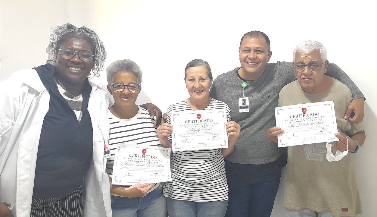 A imagem mostra um grupo de seis pessoas sorrindo diante de uma parede branca. Três delas — duas mulheres e um homem — seguram certificados de conclusão do Curso de Inclusão Digital da UBS Horto Florestal. Os certificados estão impressos em papel branco com detalhes em preto e vermelho. À esquerda da imagem, uma mulher com jaleco branco e cabelos grisalhos está sorrindo, aparentando ser uma das responsáveis pelo curso ou profissional da unidade. À direita, um homem com crachá e outro com cabelo branco completam o grupo, todos visivelmente felizes pela conquista. A imagem transmite um clima de celebração, inclusão e valorização da aprendizagem digital entre pessoas adultas e idosas.