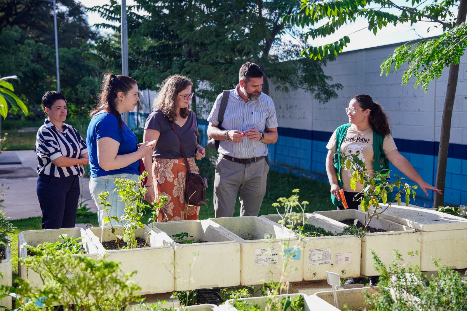 Em área externa arborizada, cinco pessoas estão reunidas ao lado de canteiros elevados com hortaliças e plantas cultivadas. Uma mulher de colete verde aponta para uma das caixas de cultivo enquanto conversa com o grupo. Ao fundo, há árvores e um muro branco e azul, sob luz natural do dia