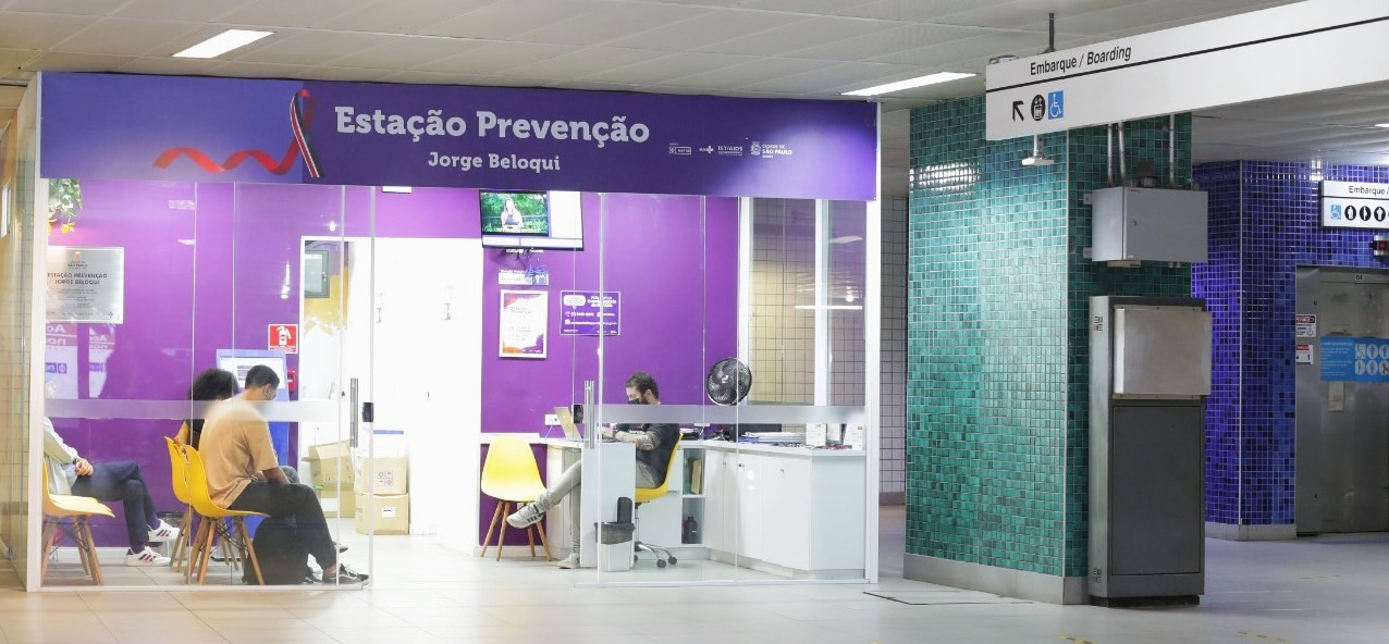 A imagem mostra a Estação Prevenção Jorge Beloqui, localizada dentro de uma estação de metrô. O espaço tem paredes de vidro e uma identidade visual marcante na cor roxa. No letreiro acima da entrada, vê-se o nome da unidade acompanhado de uma fita vermelha, símbolo da luta contra o HIV/AIDS. No interior da sala, há algumas pessoas sentadas em cadeiras amarelas, aguardando atendimento. Um profissional trabalha em uma mesa, também em uma cadeira amarela, com computador e outros materiais. A sala é iluminada, organizada e acolhedora, transmitindo um ambiente de saúde e cuidado. À direita da imagem, vê-se parte da estação de metrô com sinalização de “embarque” em português e inglês, além de painéis de azulejos em tons de verde e azul, característicos da arquitetura de estações de transporte público em São Paulo. A Estação Prevenção é uma iniciativa voltada à promoção da saúde e prevenção de infecções sexualmente transmissíveis, homenageando Jorge Beloqui, ativista dos direitos das pessoas vivendo com HIV/AIDS