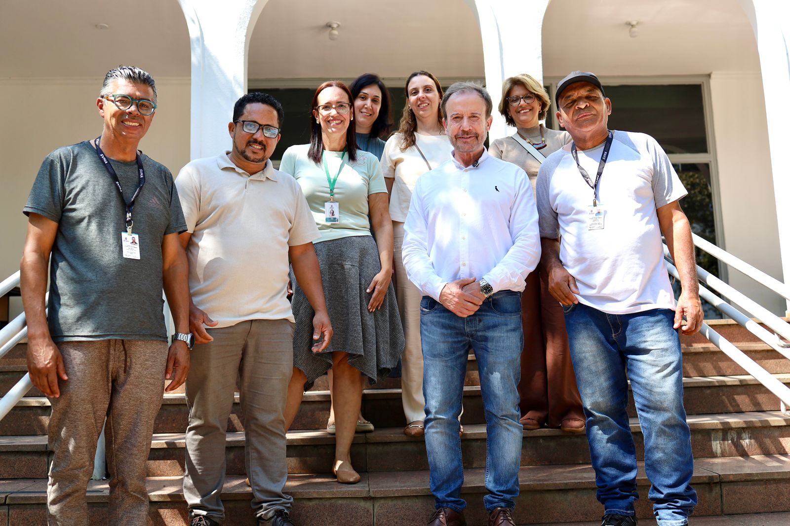 A foto mostra um grupo de sete pessoas reunidas em uma escadaria, posando para uma fotografia em frente a um prédio de fachada clara, com grandes arcos e janelas ao fundo. Na primeira fila, há quatro pessoas. Duas delas usam crachás pendurados no pescoço, e todas estão olhando para a câmera. Ao centro, um homem de camisa branca de manga longa e jeans está com as mãos entrelaçadas à frente do corpo. À direita, um homem usando boné, camiseta branca e jeans também aparece com um crachá. Na segunda fila, há três mulheres, posicionadas um pouco acima na escadaria. Elas estão sorrindo e usando roupas variadas, incluindo blusas claras e uma saia estampada. Algumas pessoas usam crachás de identificação, sugerindo que o grupo pode estar relacionado a um evento, instituição ou atividade profissional. 