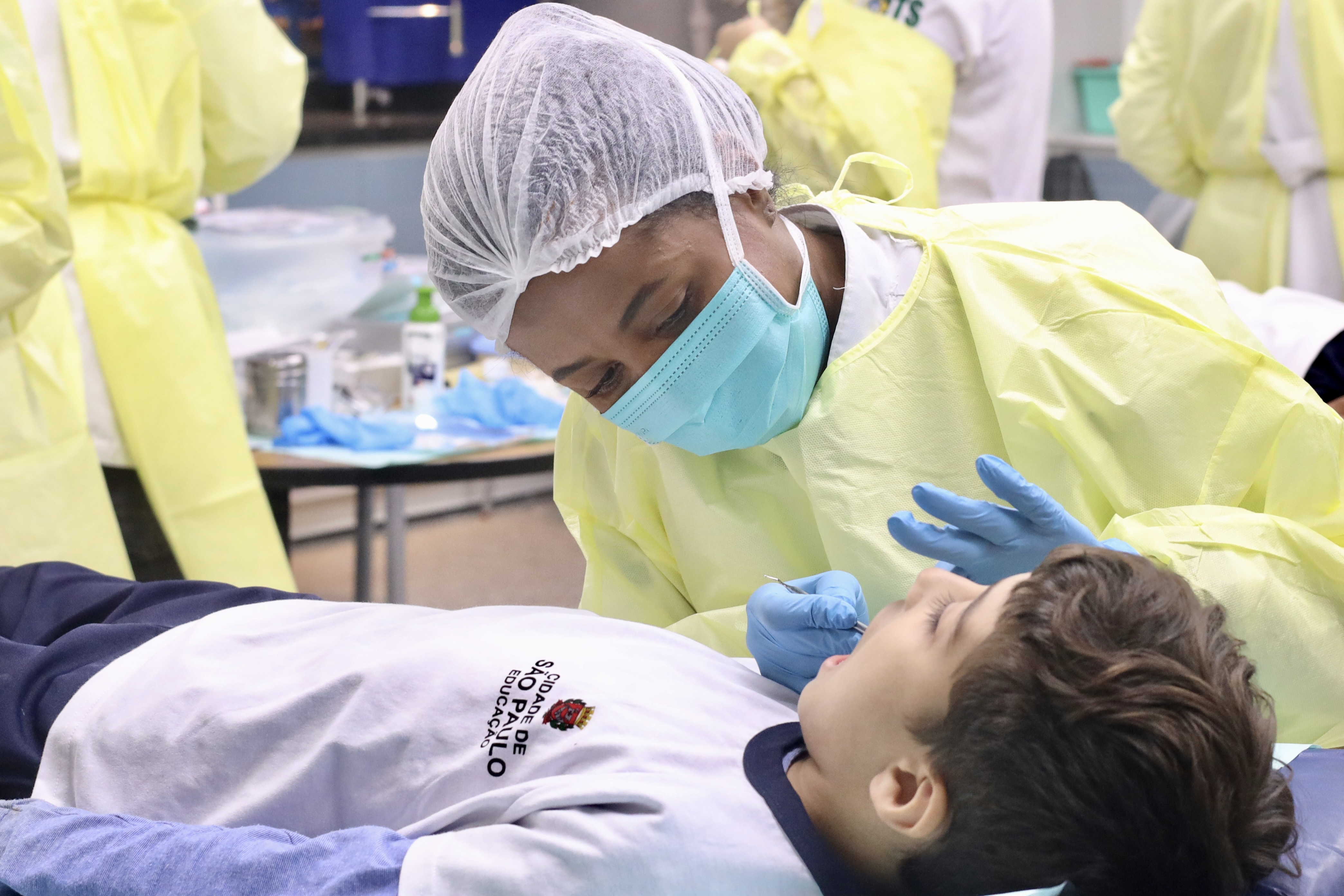 A imagem mostra uma profissional de saúde realizando um atendimento odontológico em uma criança. A profissional, uma mulher negra, está inclinada sobre o paciente e veste equipamentos de proteção individual (EPIs), incluindo um capote cirúrgico amarelo, máscara descartável azul-clara, touca protetora branca e luvas azuis. Ela manuseia um instrumento cirúrgico/odontológico próximo à boca da criança com cuidado.   A criança está deitada de costas e veste o uniforme escolar da rede municipal de ensino, composto por uma camiseta branca com o brasão e a inscrição 
