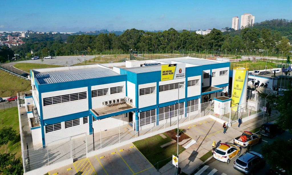 A imagem mostra a fachada de uma unidade de saúde pública da cidade de São Paulo, identificada como UBS Helena Miguel Rezek. O prédio é de dois andares, com pintura nas cores azul, branco e cinza, e apresenta uma estrutura moderna e funcional. Na parte frontal, há placas com o nome da unidade e o logotipo da Prefeitura de São Paulo. À direita da construção, destaca-se uma torre cilíndrica amarela com informações institucionais, incluindo o nome da unidade e referências à rede municipal de saúde. Em frente ao prédio, há uma área cercada e pavimentada, com espaço de acesso e estacionamento. Ao fundo, é possível ver árvores, áreas verdes e diversos prédios residenciais ou comerciais, indicando que a unidade está situada em uma região urbana. O céu está claro e azul, sugerindo um dia ensolarado.