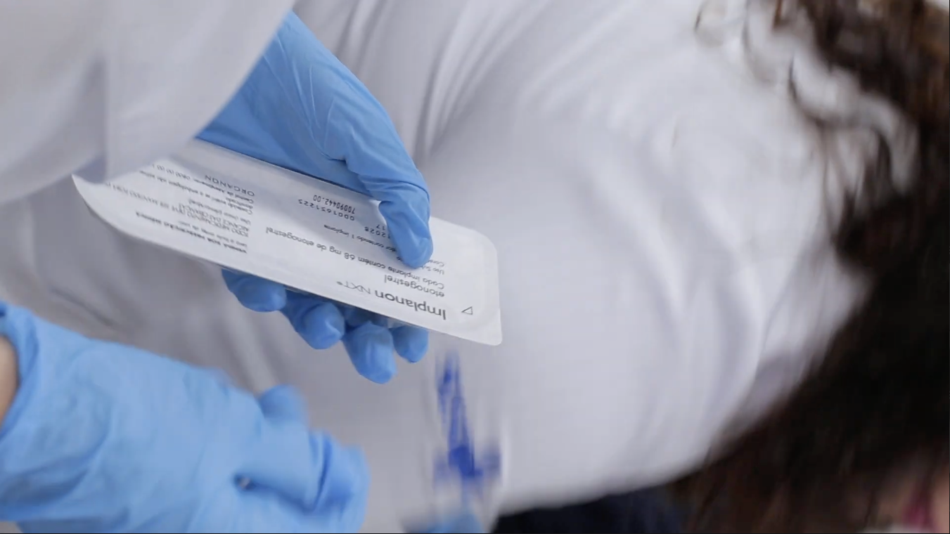 A imagem mostra um close de um atendimento de saúde: uma profissional com luvas azuis segura uma embalagem de medicamento — é possível ler “Implanon NXT”, um implante contraceptivo subdérmico. Ao fundo, aparece parte do corpo de outra pessoa vestida com roupa branca, em uma cena que sugere orientação ou preparo para um procedimento clínico.