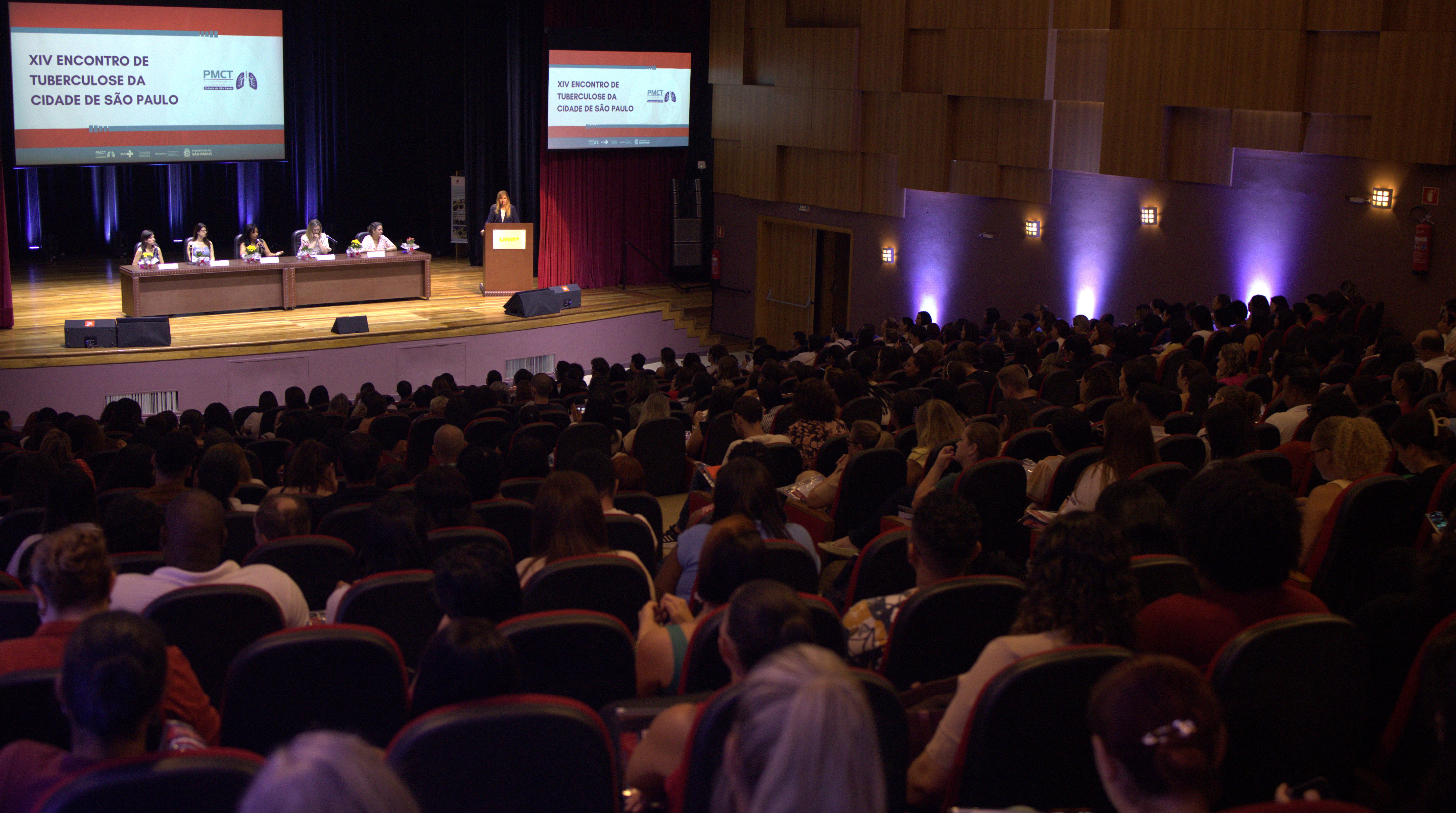 A imagem mostra um auditório amplo e lotado durante um evento institucional na área da saúde. O público ocupa praticamente todas as poltronas, voltado para o palco, onde ocorre uma mesa de abertura com cinco participantes sentados e uma pessoa discursando em um púlpito. No fundo do palco, dois telões exibem o título do evento: “XIV Encontro de Tuberculose da Cidade de São Paulo”, acompanhado de logotipos institucionais. A iluminação cênica em tons de roxo e azul valoriza o ambiente, que tem acabamento em madeira nas paredes laterais, conferindo um aspecto formal e organizado.