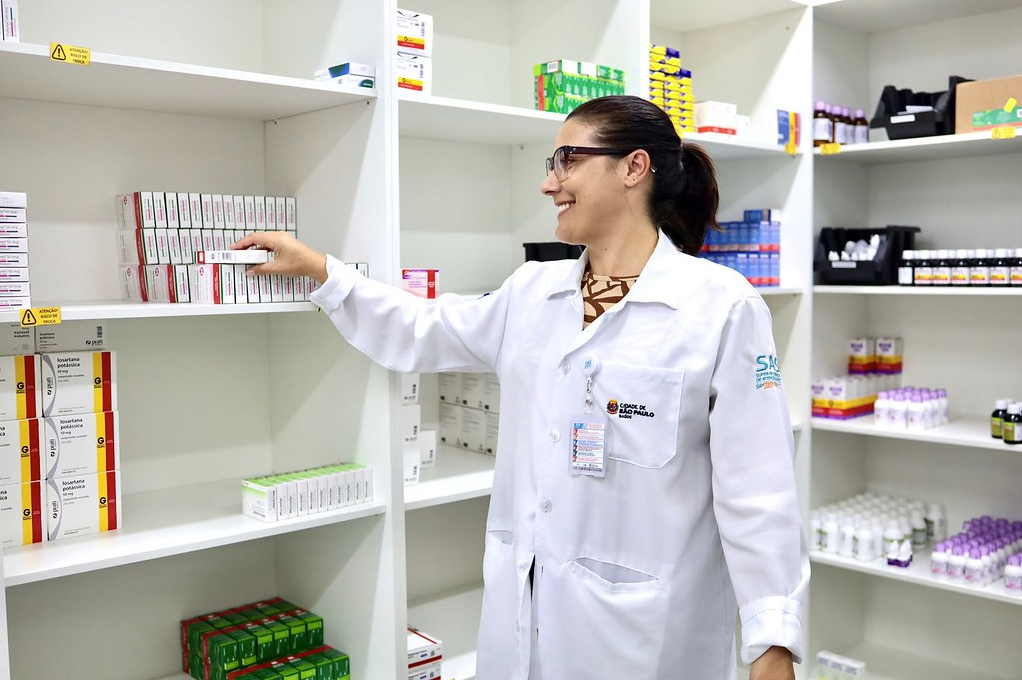 A imagem mostra uma profissional de saúde dentro da farmácia da UBS Engenheiro Trindade. Ela veste jaleco branco com o crachá da Prefeitura de São Paulo e óculos, e aparece sorrindo enquanto retira uma caixa de medicamento de uma das prateleiras.  O ambiente é claro, organizado e bem iluminado. As estantes brancas ao fundo estão repletas de caixas de remédios e frascos, todos dispostos de forma ordenada, com identificação visível. É possível ver diferentes tipos de embalagens, indicando a variedade de medicamentos disponíveis para atendimento à população.