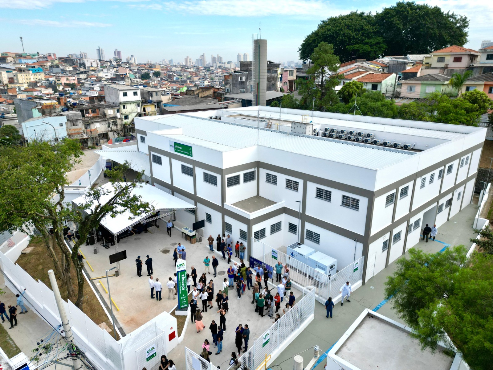 A foto revela o dia da inauguração da Unidade de Pronto-Atendimento(UPA)Ipiranga. Muitas pessoas estão circulando no pátio de entrada no equipamento de saúde.  Uma placa a esquerde nas cores verde e branca  destacam o nome do equipamento de saúde, UPA Ipiranga, e ao fundo o prédio. A estrutura dele foi pintada na cor branca. À esquerda da entrada da unidade de saúde tem uma árvore. A foto tirada do alto mostra a amplitude do equipamento.   
