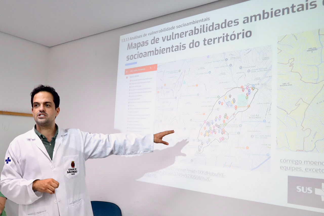 Thiago Menezes, gerente da UBS Jardim Comercial, de jaleco branco, apresenta o biomapa do território. Ao fundo, uma projeção mostra mapas de vulnerabilidades socioambientais, enquanto ele aponta para uma das áreas mapeadas.