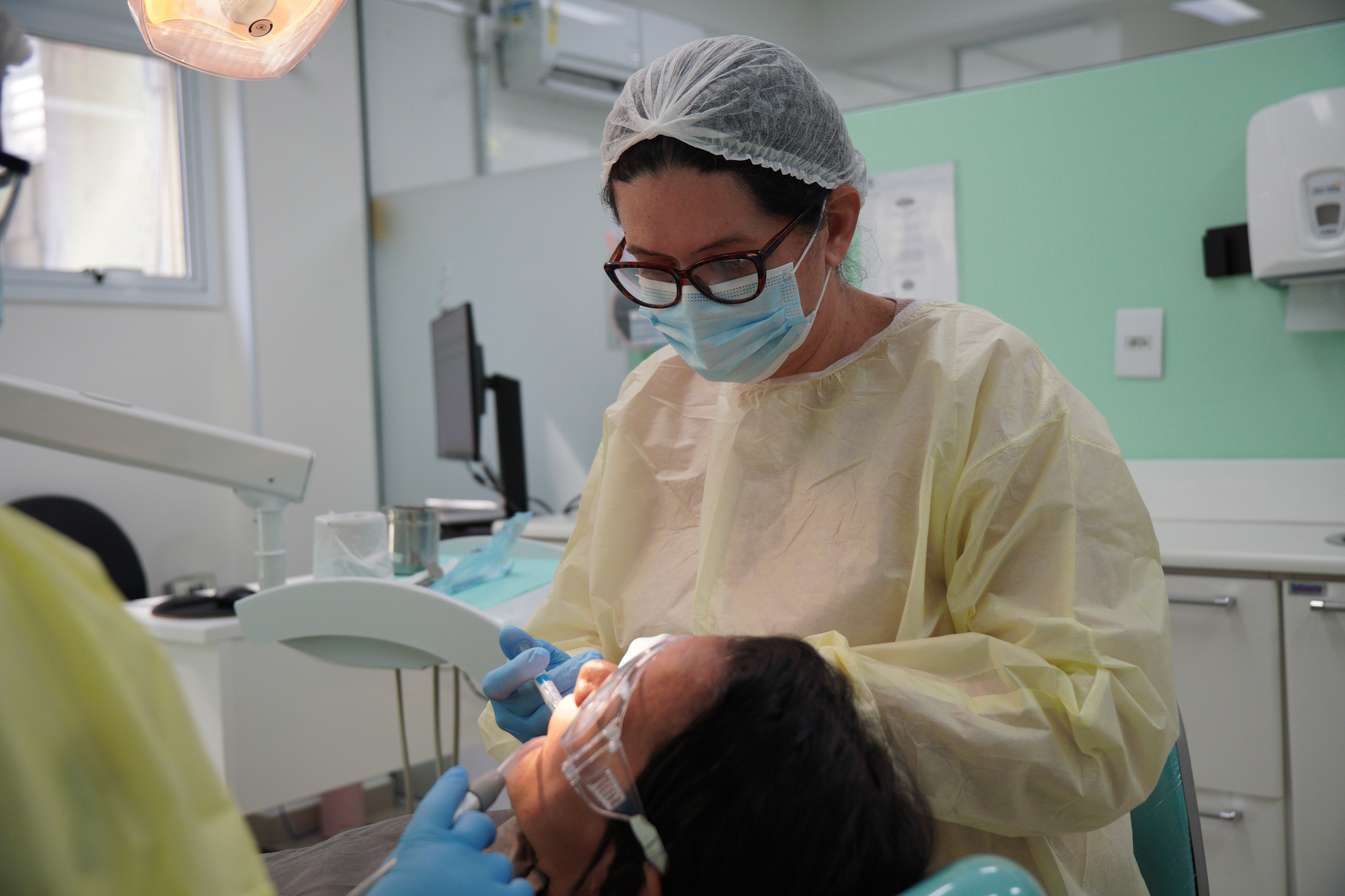 Profissional de saúde realiza atendimento odontológico em paciente, em ambiente clínico, utilizando equipamentos de proteção individual, como máscara, touca, avental e luvas. A cena representa o atendimento especializado e os cuidados oferecidos na rede pública de saúde bucal.