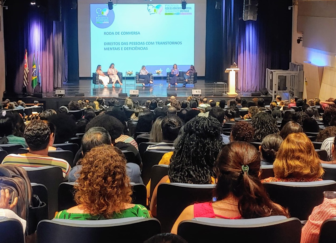 A foto mostra um auditório lotado, visto a partir dos fundos; no palco, à frente, em sofás, estão sentadas cinco mulheres, conversando; atrás delas, em um telão, lê-se: “Roda de conversa – direitos das pessoas com transtornos mentais e deficiências