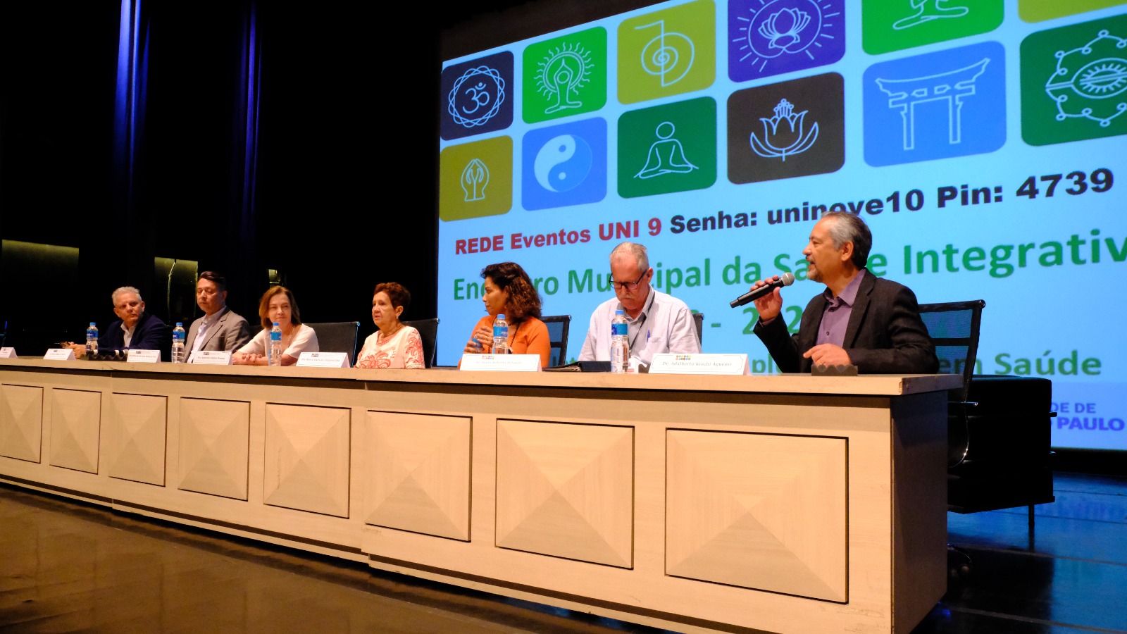 A foto mostra uma mesa composta por sete pessoas homens e mulheres. Um integrante fala ao microfone do lado direito. Ao fundo há um painel com vários ícones e, abaixo, a inscrição do evento Encontro Municipal da Saúde Integrativa.  