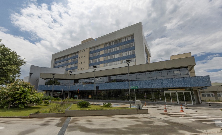 A foto mostra a fachada de um prédio com cerca de cinco andares, nas cores azul e bege; em frente, uma placa indica tratar-se de um hospital