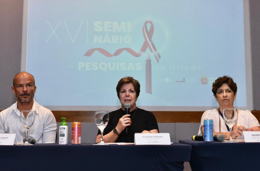 A imagem mostra três pessoas, duas mulheres e um homem, sentadas em uma mesa de um evento, com um fundo de projeção que exibe o título