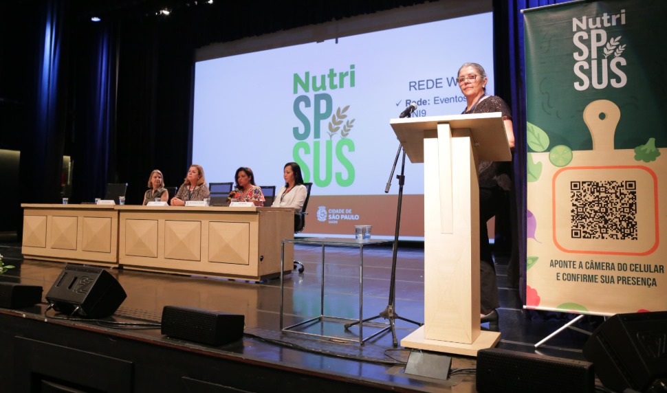 A imagem mostra um evento em um auditório, com cinco pessoas sentadas em uma mesa sobre um palco e uma mulher em pé discursando em um púlpito à direita. O fundo apresenta um telão exibindo o logotipo