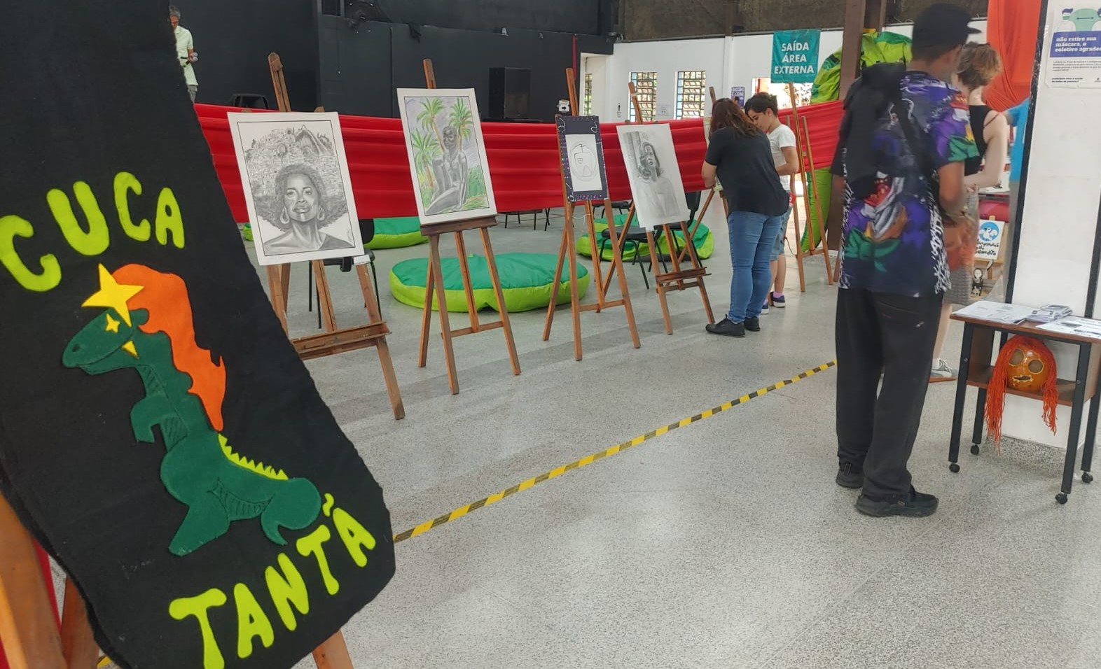 A imagem mostra uma exposição de arte em um ambiente interno. Há diversos cavaletes alinhados em fila, cada um exibindo desenhos ou pinturas com temáticas variadas, incluindo retratos e paisagens. No lado esquerdo, um banner ou painel preto apresenta um desenho estilizado de uma figura que remete à