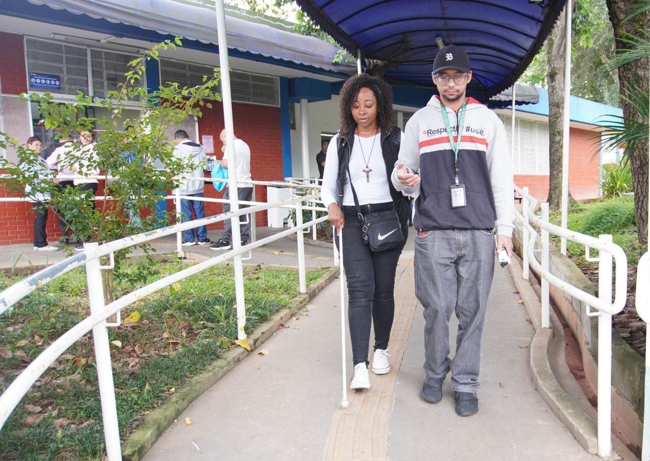 No fundo as pessoas estão interagindo ao atendimento no Centro Especializado em Reabilitação(CER) e, descendo a rampa, estão dois pacientes com deficiência visual. A mulher está com a bengala com ponteira branca e o homem está ao seu lado.