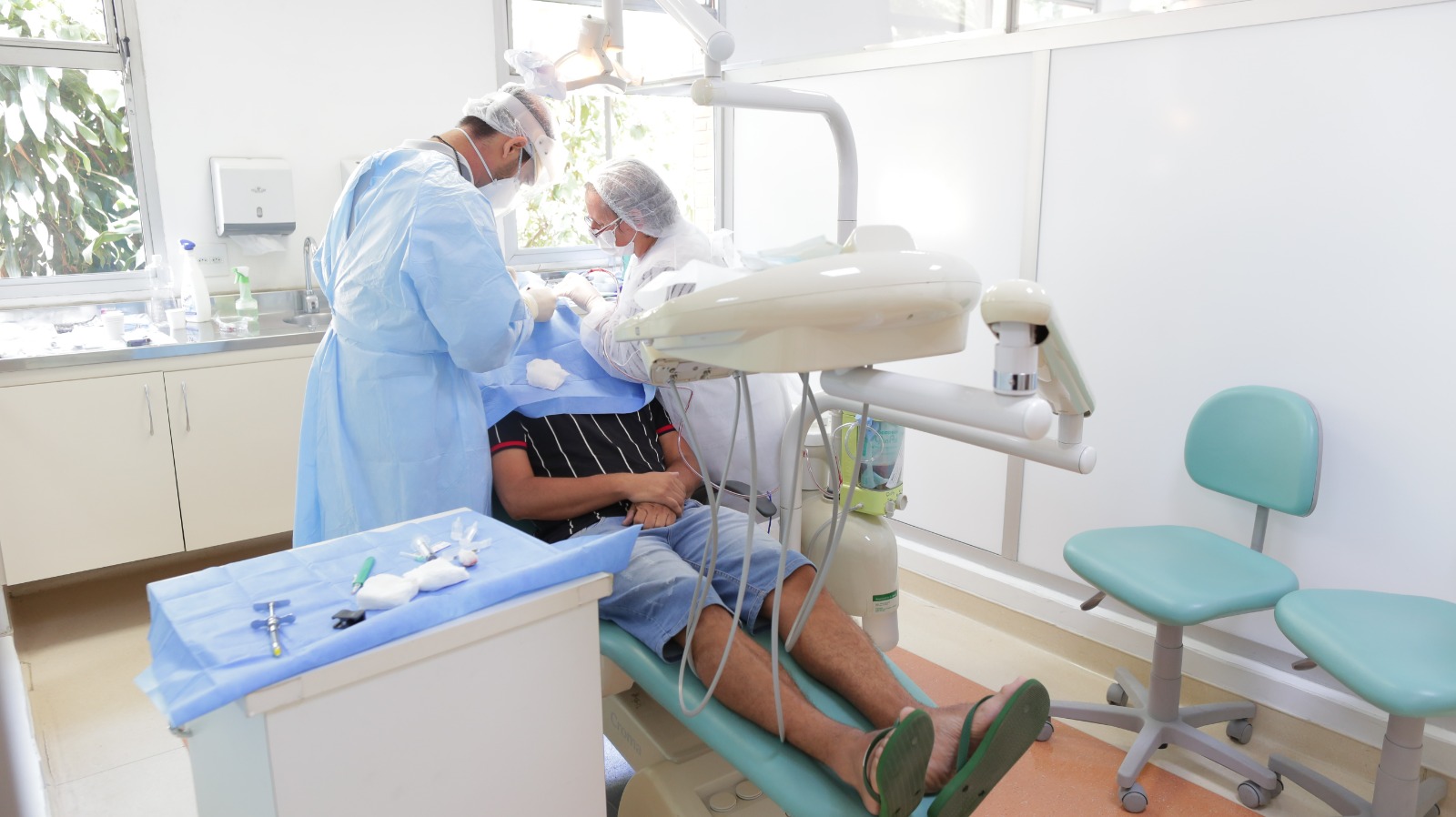 A imagem mostra uma consulta odontológica em andamento. O paciente está deitado em uma cadeira odontológica, vestindo bermuda jeans e camiseta listrada, com os pés calçados em chinelos. Dois profissionais de saúde, usando jalecos, toucas, máscaras e luvas, trabalham no atendimento do paciente. Um deles está à esquerda, de costas, e o outro está ao lado do paciente, mexendo no procedimento. O consultório apresenta um ambiente limpo e bem iluminado, com paredes brancas e uma ampla janela, que permite a entrada de luz natural. Há equipamentos odontológicos tradicionais, como a cadeira, um refletor e instrumentos cirúrgicos organizados sobre uma bandeja coberta por um tecido azul.