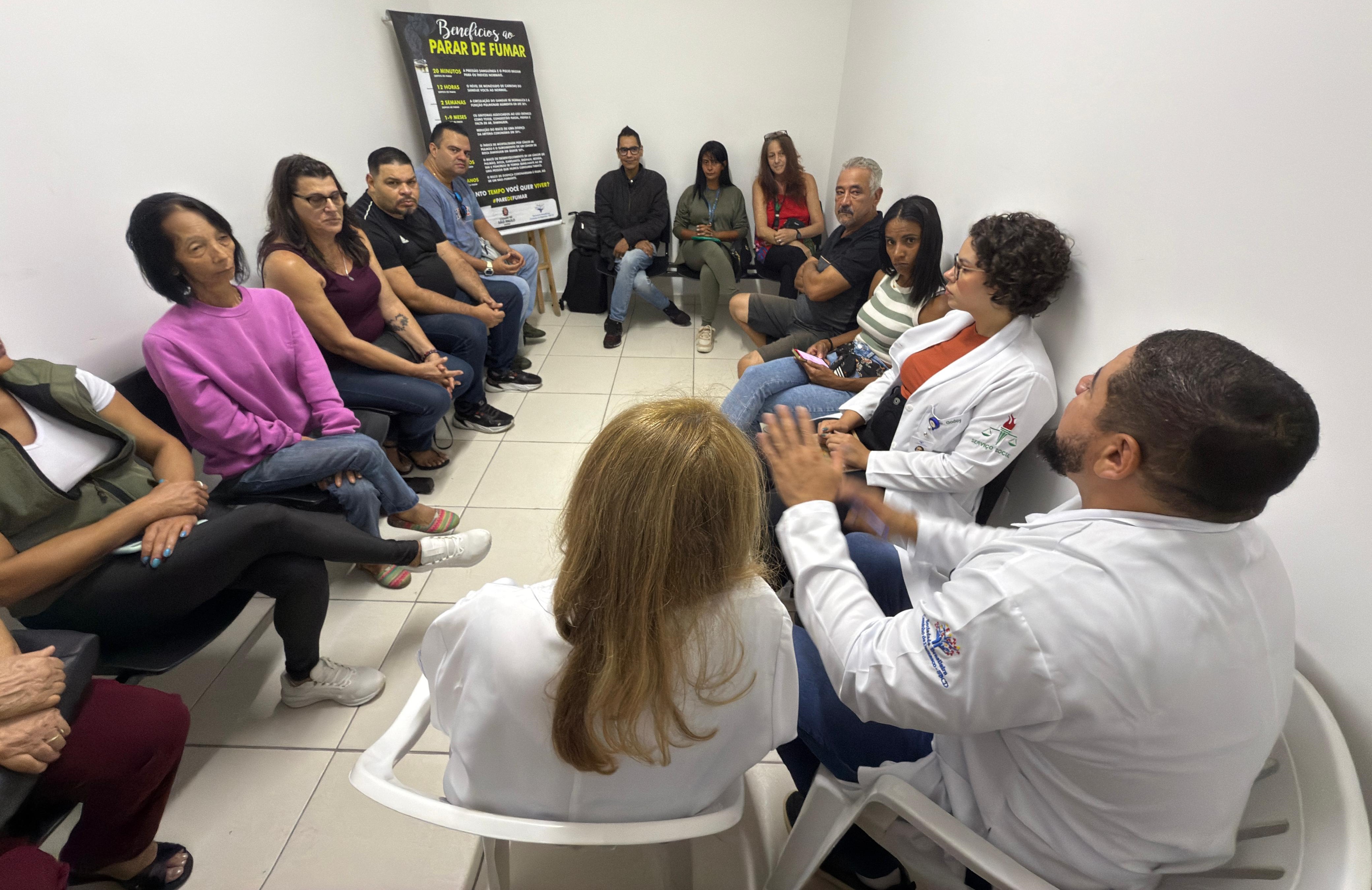A imagem mostra uma roda de conversa em uma sala branca, onde cerca de vinte pessoas estão sentadas em círculo. Entre os participantes, há profissionais da saúde — identificáveis pelos jalecos brancos — e usuários do serviço de saúde, o que indica um ambiente de acolhimento e orientação. A atmosfera parece ser de escuta e troca de experiências. No centro da imagem, um dos profissionais de jaleco gesticula enquanto fala, sugerindo que está conduzindo a reunião ou realizando uma intervenção. Na parede ao fundo, há um cartaz com o título “Benefícios de Parar de Fumar”, indicando que o encontro está relacionado a um grupo de apoio para cessação do tabagismo. O cenário sugere uma ação educativa e de promoção da saúde, voltada para o cuidado com pessoas que desejam parar de fumar.
