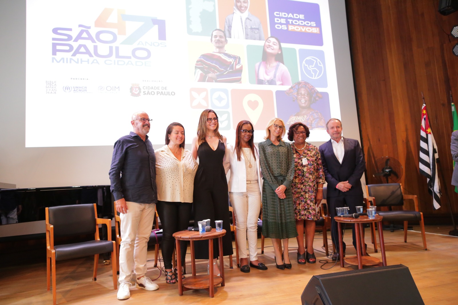 A imagem mostra um grupo de sete pessoas posando juntas em um palco, em frente a um telão com o texto “471 anos São Paulo, minha cidade” e o slogan “Cidade de todos os povos!” no canto superior direito. O telão também exibe logotipos de organizações parceiras, como a ACNUR, OIM e a Prefeitura de São Paulo, além de fotos de diversas pessoas representando a diversidade cultural. As pessoas no palco estão alinhadas lado a lado, sorrindo para a câmera. O ambiente é iluminado e possui cadeiras e mesas no palco. No lado direito, há bandeiras de São Paulo e do Brasil.