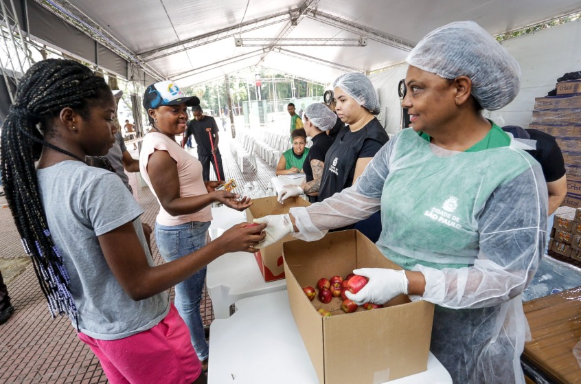 A imagem mostra uma ação de distribuição de alimentos em um evento organizado pela Prefeitura de São Paulo. Sob uma grande tenda, voluntárias vestindo aventais, toucas e luvas de proteção distribuem frutas para as pessoas que estão na fila. No primeiro plano, uma mulher voluntária segura uma maçã e entrega para uma jovem de camiseta cinza e calça rosa. Atrás dela, outra mulher de boné azul e camiseta rosa aguarda na fila, segurando um pacote de alimento. Ao fundo, há mais voluntários e pessoas organizando a distribuição, além de caixas de suprimentos empilhadas