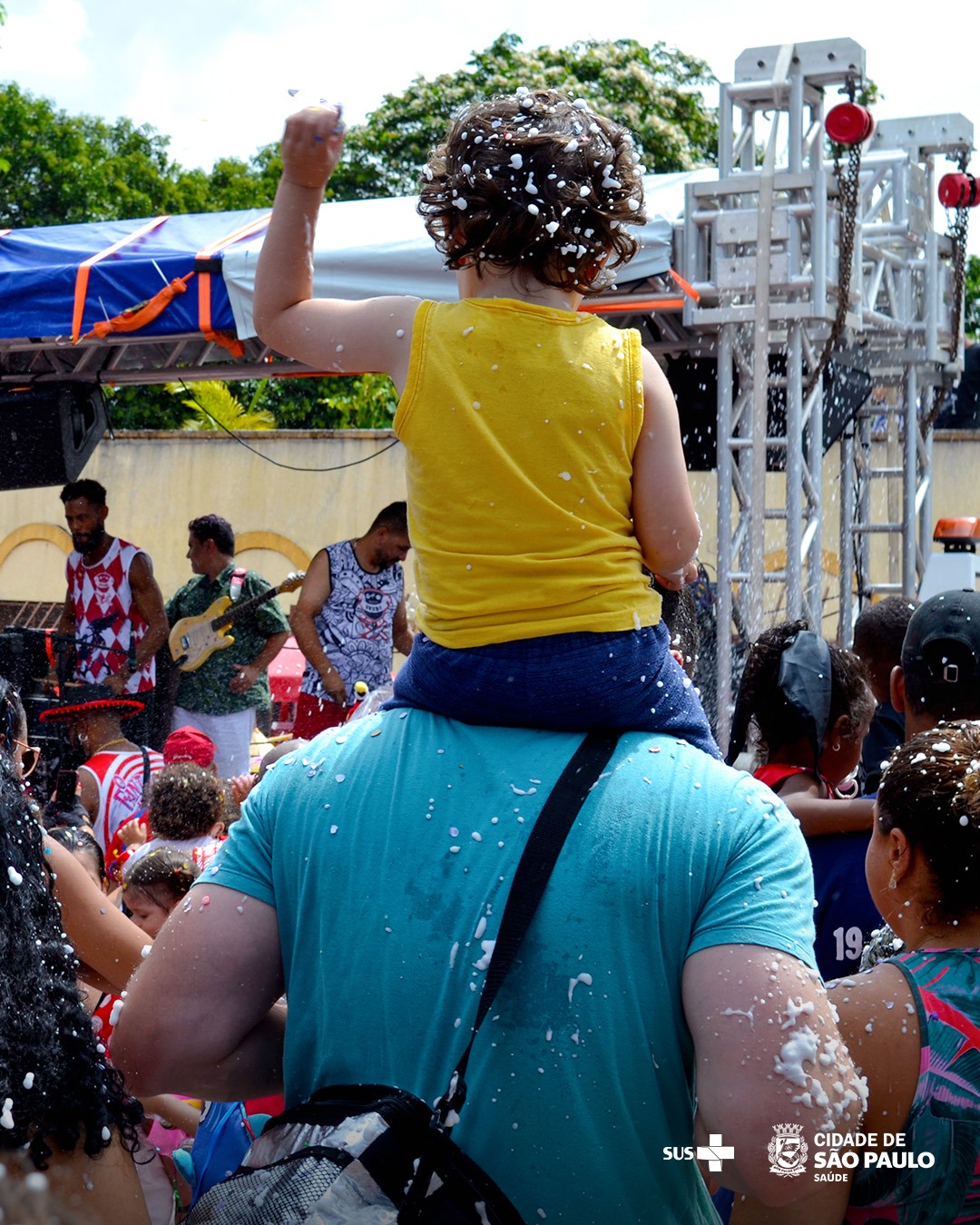 A imagem retrata uma cena animada de Carnaval de rua, com um adulto vestindo uma camiseta azul carregando uma criança nos ombros. A criança, de cabelos cacheados, veste uma blusa amarela e levanta o braço, brincando com espuma, que está espalhada por várias pessoas ao redor. No fundo, há um palco com músicos tocando instrumentos, e o ambiente está repleto de foliões vestidos com roupas coloridas e fantasias. A presença do logotipo da