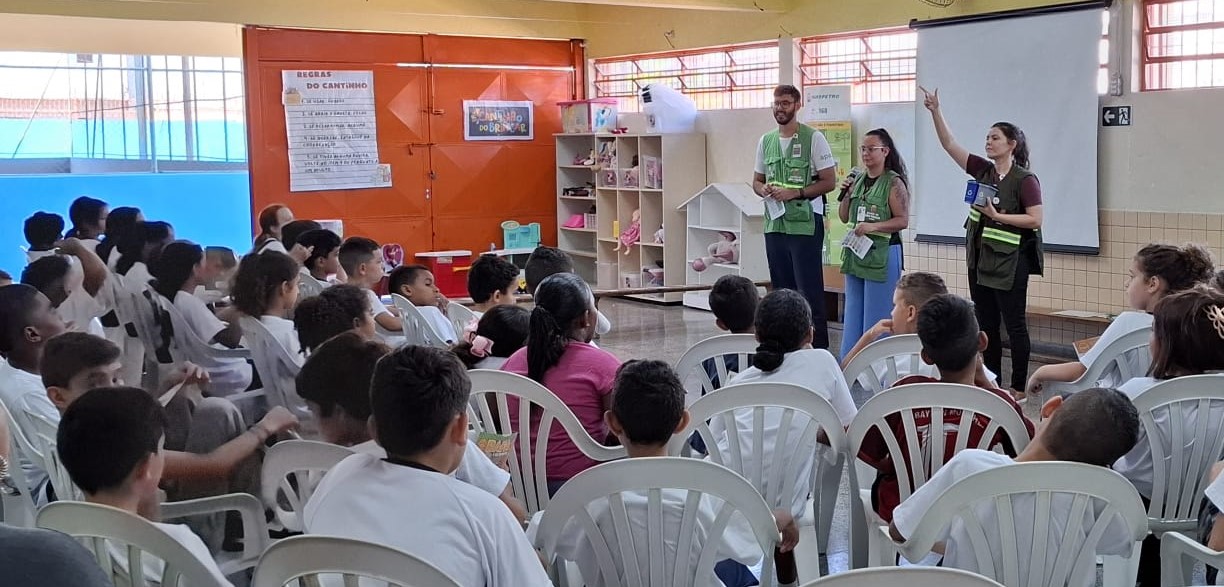 A imagem mostra uma atividade educativa em um ambiente escolar, onde um grupo de crianças vestindo uniformes escolares está sentado em cadeiras brancas de plástico, assistindo atentamente a uma apresentação. Três adultos, usando coletes verdes identificadores, estão à frente da sala conduzindo a atividade. Uma das mulheres está segurando um microfone e falando, enquanto a outra gesticula com a mão levantada, segurando um objeto azul. O terceiro adulto, um homem, segura alguns panfletos.  O ambiente é iluminado e organizado, com prateleiras contendo brinquedos e materiais educativos ao fundo. Há também um quadro de projeção e um cartaz na parede com regras do 