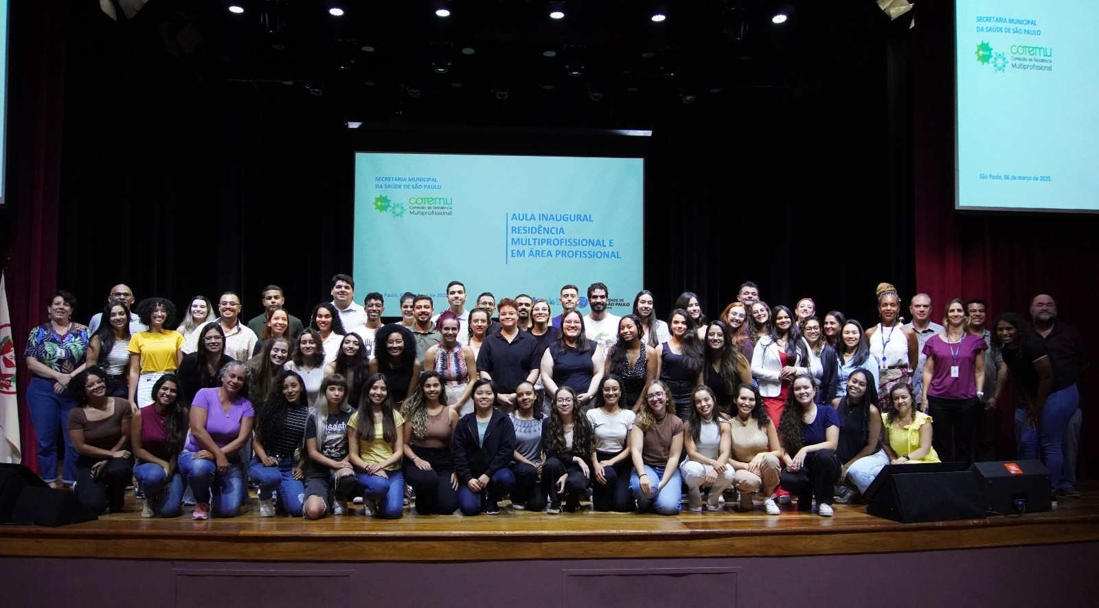 A imagem mostra um grande grupo de pessoas reunidas em um auditório para um evento oficial. No centro do palco, há um telão projetando o título