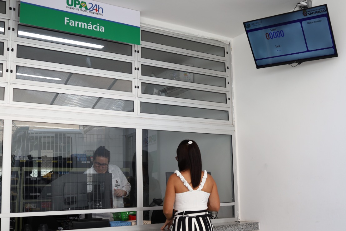 A imagem mostra o balcão de uma farmácia localizada dentro de uma unidade de pronto atendimento (UPA 24h). Uma mulher de costas, vestindo uma blusa branca com detalhes ondulados nas alças e uma saia listrada em preto e branco, está sendo atendida por uma funcionária que veste jaleco branco e está atrás de um vidro, utilizando um computador. Acima do balcão, há uma placa indicando