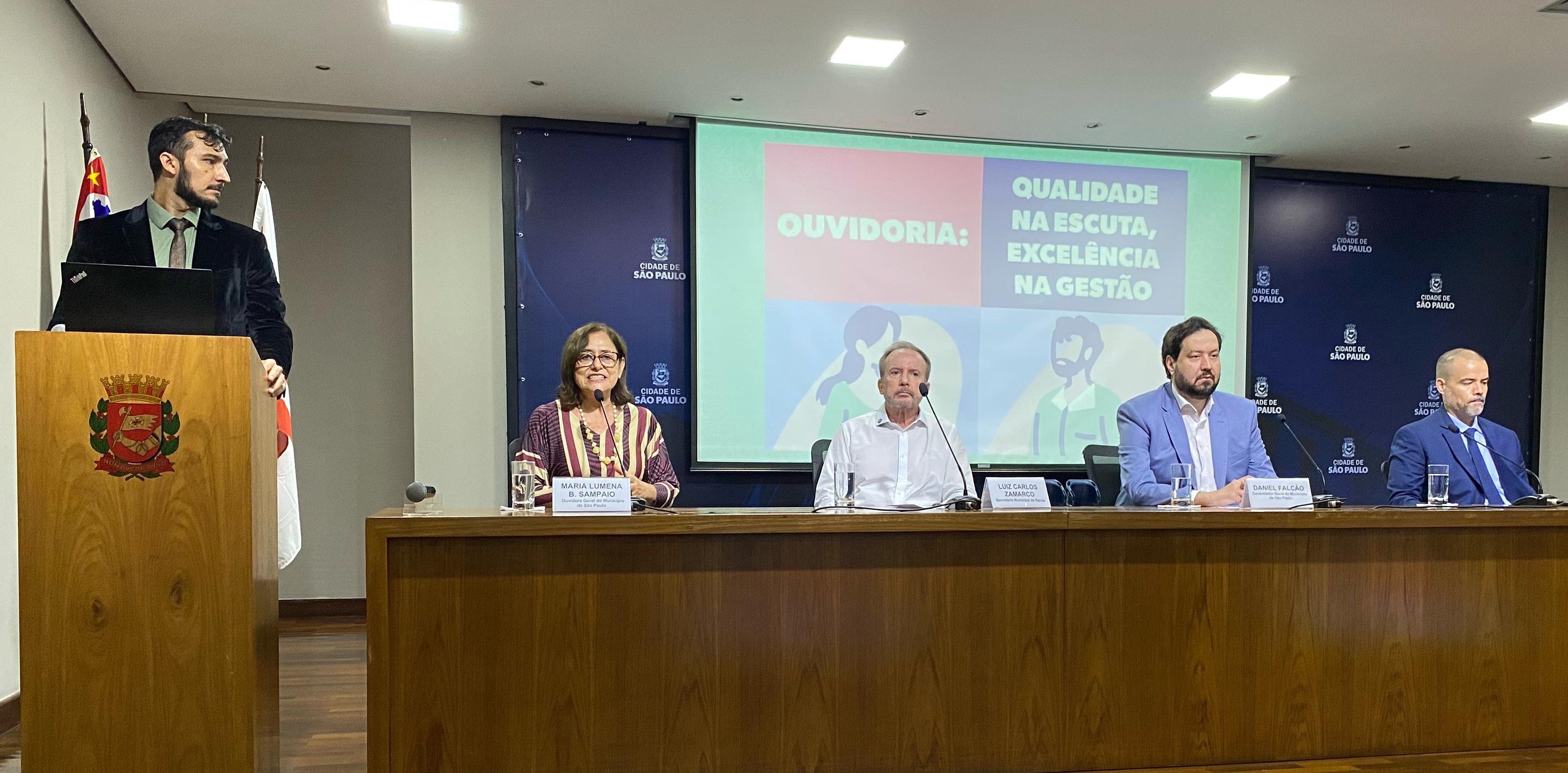 A imagem mostra um evento oficial realizado na cidade de São Paulo, com quatro pessoas sentadas em uma mesa e um palestrante de pé em um púlpito de madeira. O painel de fundo tem a logomarca da cidade de São Paulo e exibe a frase 