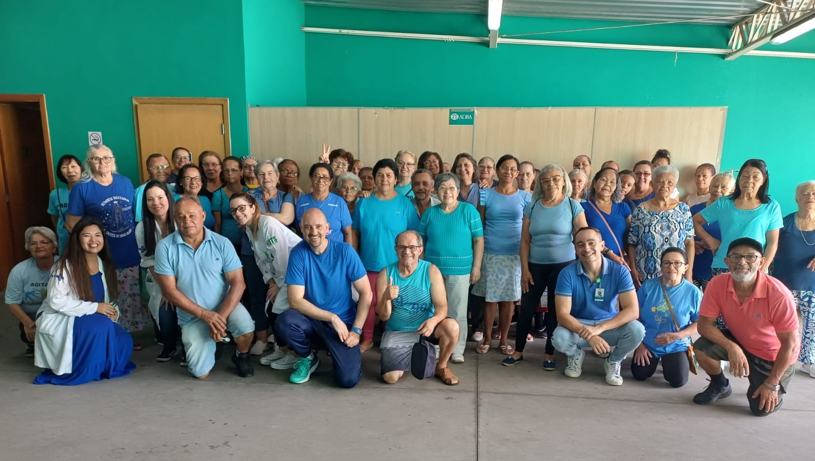 A imagem mostra um grupo grande de pessoas, predominantemente idosas, em um ambiente interno com paredes verdes. Eles estão usando roupas na tonalidade azul, com algumas variações no estilo e tom da cor. No centro, há dois homens agachados em primeiro plano, sorrindo para a câmera. Ao redor deles, há várias outras pessoas de pé, algumas com expressões sorridentes e outras fazendo gestos divertidos, como o sinal de paz e amor. O ambiente parece ser um espaço dedicado a atividades para os idosos, possivelmente uma atividade física. 