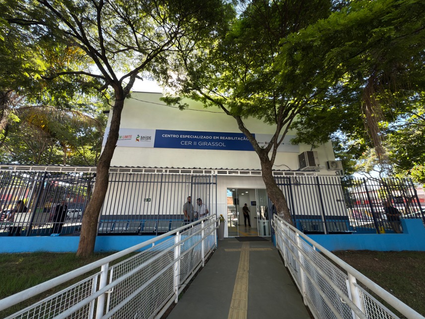 A imagem mostra a fachada do Centro Especializado em Reabilitação CER II Girassol, localizado em uma área arborizada. A entrada principal do prédio é acessível, com uma rampa adaptada com corrimãos laterais e piso tátil, sinalizando um compromisso com a acessibilidade para pessoas com deficiência.  O prédio é de cor clara e tem uma placa azul no topo com os dizeres: 