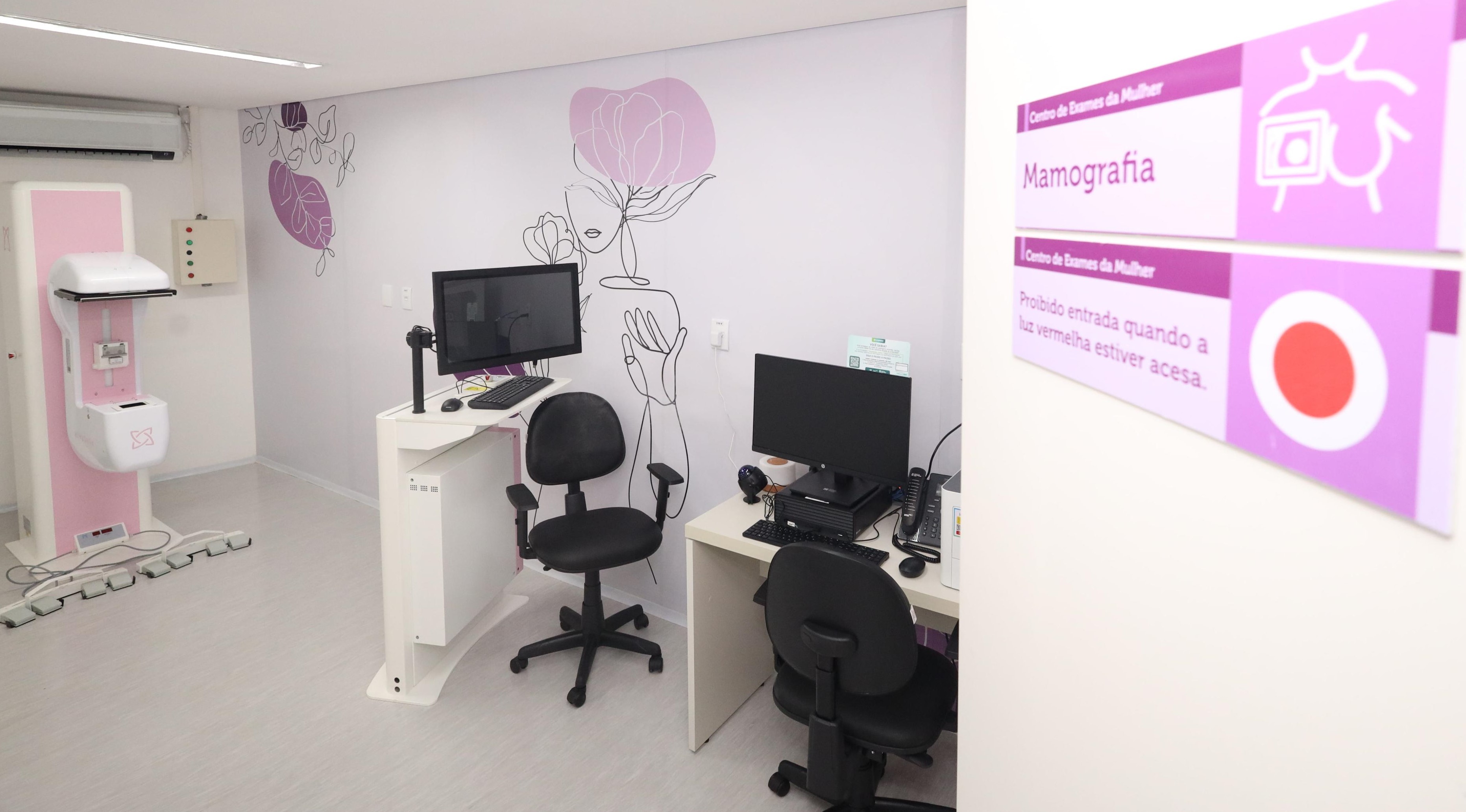 A imagem mostra uma sala de mamografia, parte de um Centro de Exames da Mulher. O ambiente é bem iluminado, com paredes brancas e decoração em tons de rosa e lilás, remetendo ao cuidado com a saúde feminina. Há um mural com uma ilustração estilizada de uma mulher com uma flor, trazendo um toque acolhedor e artístico ao espaço. Na parede à direita, há uma placa informativa com os dizeres: Centro de Exames da Mulher Mamografia “Proibido entrada quando a luz vermelha estiver acesa” A sala conta com um equipamento de mamografia à esquerda, de cor branca com detalhes em rosa. Também há duas estações de trabalho com computadores e cadeiras, possivelmente para técnicos ou médicos que operam os exames e analisam as imagens. O ambiente está limpo, organizado e acessível, transmitindo profissionalismo e acolhimento ao mesmo tempo. É um espaço voltado ao diagnóstico precoce e cuidado da saúde da mulher, reforçado pela paleta de cores suave e elementos visuais delicados