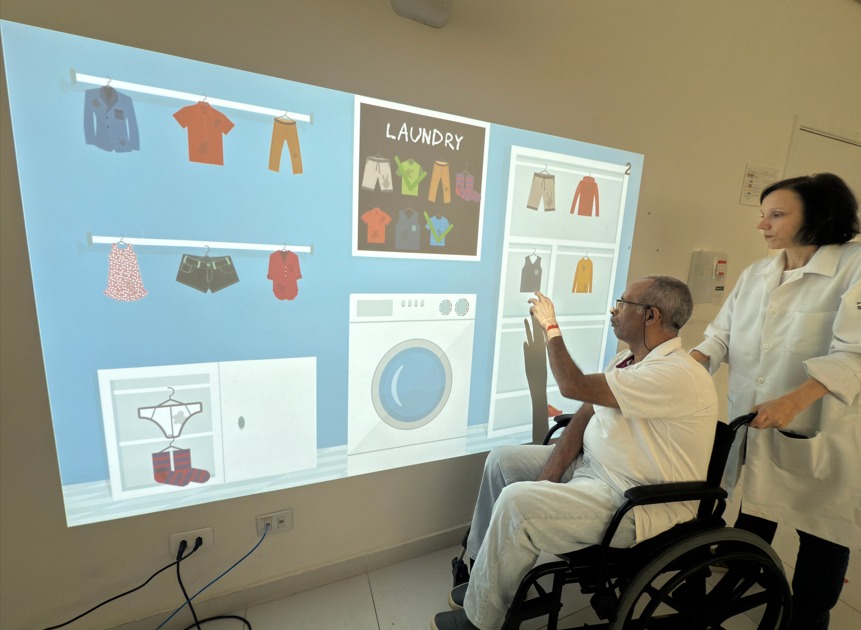 A imagem mostra uma cena de reabilitação ou terapia ocupacional com o uso de tecnologia interativa. Um homem em cadeira de rodas está interagindo com uma parede onde é projetada uma imagem digital colorida com o tema de lavanderia (laundry). Ele toca com a mão a projeção, que inclui ilustrações de roupas penduradas, uma máquina de lavar e armários. Ao lado dele, uma profissional de saúde — possivelmente uma terapeuta ocupacional ou fisioterapeuta — observa e o acompanha atentamente. Ambos estão vestindo jalecos brancos, o que indica um ambiente clínico ou hospitalar. A terapeuta segura a cadeira de rodas, demonstrando apoio ao paciente durante a atividade. A projeção parece ser parte de um sistema de reabilitação digital que estimula funções cognitivas e motoras, por meio de atividades do cotidiano em formato interativo. A cena transmite cuidado, inovação tecnológica na saúde e humanização no atendimento