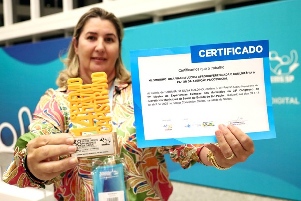 A imagem mostra uma mulher segurando um certificado e um troféu, sorrindo discretamente para a câmera. O certificado reconhece o trabalho intitulado