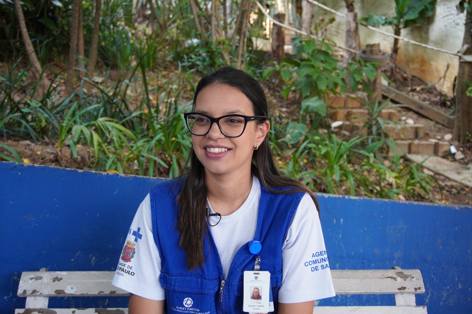 A imagem mostra Camila, uma mulher jovem sentada em um banco de madeira branco, em um ambiente ao ar livre cercado por vegetação. Ela tem cabelos castanhos escuros, lisos e longos, pele clara e usa óculos de grau com armação preta. A mulher veste uma camiseta branca com um colete azul escuro que traz identificação da Prefeitura de São Paulo e do Hospital Albert Einstein. Presa ao colete, há uma identificação funcional com sua foto e nome, indicando que ela é agente comunitária de saúde. Ela está sorrindo levemente, com expressão tranquila e receptiva.