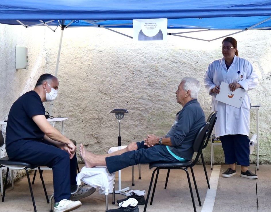 A imagem mostra uma ação de cuidado com a saúde em um ambiente ao ar livre, sob uma tenda azul. Um profissional de saúde, usando roupa escura e máscara, está examinando os pés de um idoso que está sentado em frente a ele, com as pernas estendidas sobre um apoio coberto com um lençol branco. Ao fundo, uma profissional de jaleco branco observa a consulta e segura uma prancheta. Na parede atrás da tenda, há um cartaz que indica tratar-se de uma ação relacionada ao cuidado com o “pé diabético”. A cena transmite atenção, acolhimento e cuidado especializado com a saúde da população