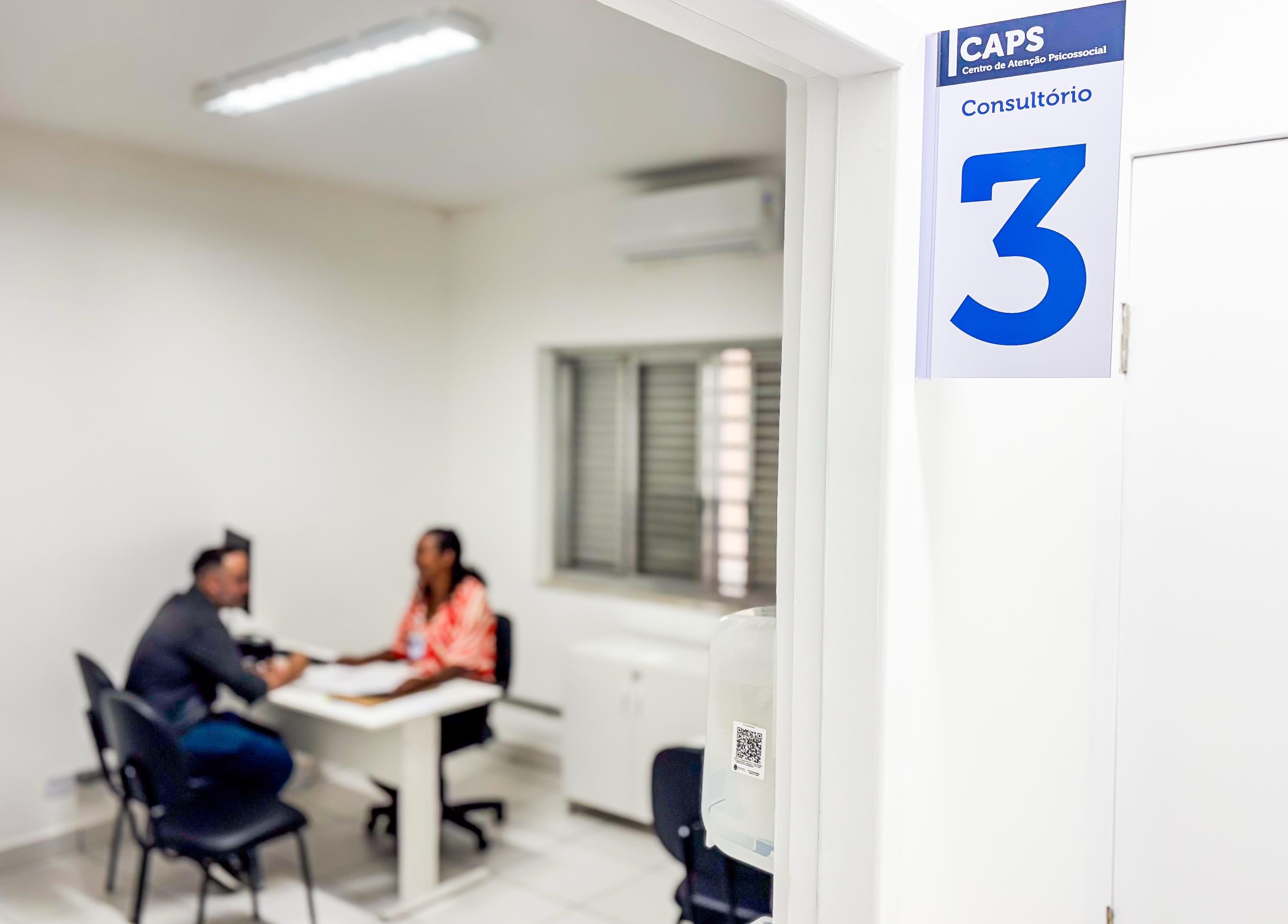 A imagem mostra o interior de um consultório no Centro de Atenção Psicossocial (CAPS), identificado como Consultório 3, conforme placa visível na entrada da sala. O ambiente é claro, limpo e bem iluminado, com paredes e mobiliário em tons claros. Dentro da sala, duas pessoas estão sentadas em lados opostos de uma mesa — um homem, de costas, conversa com uma profissional de saúde, que está usando um crachá. Embora desfocadas, ambas as pessoas parecem estar envolvidas em uma conversa atenta, sugerindo um atendimento psicossocial. A cena transmite acolhimento, escuta e cuidado em saúde mental