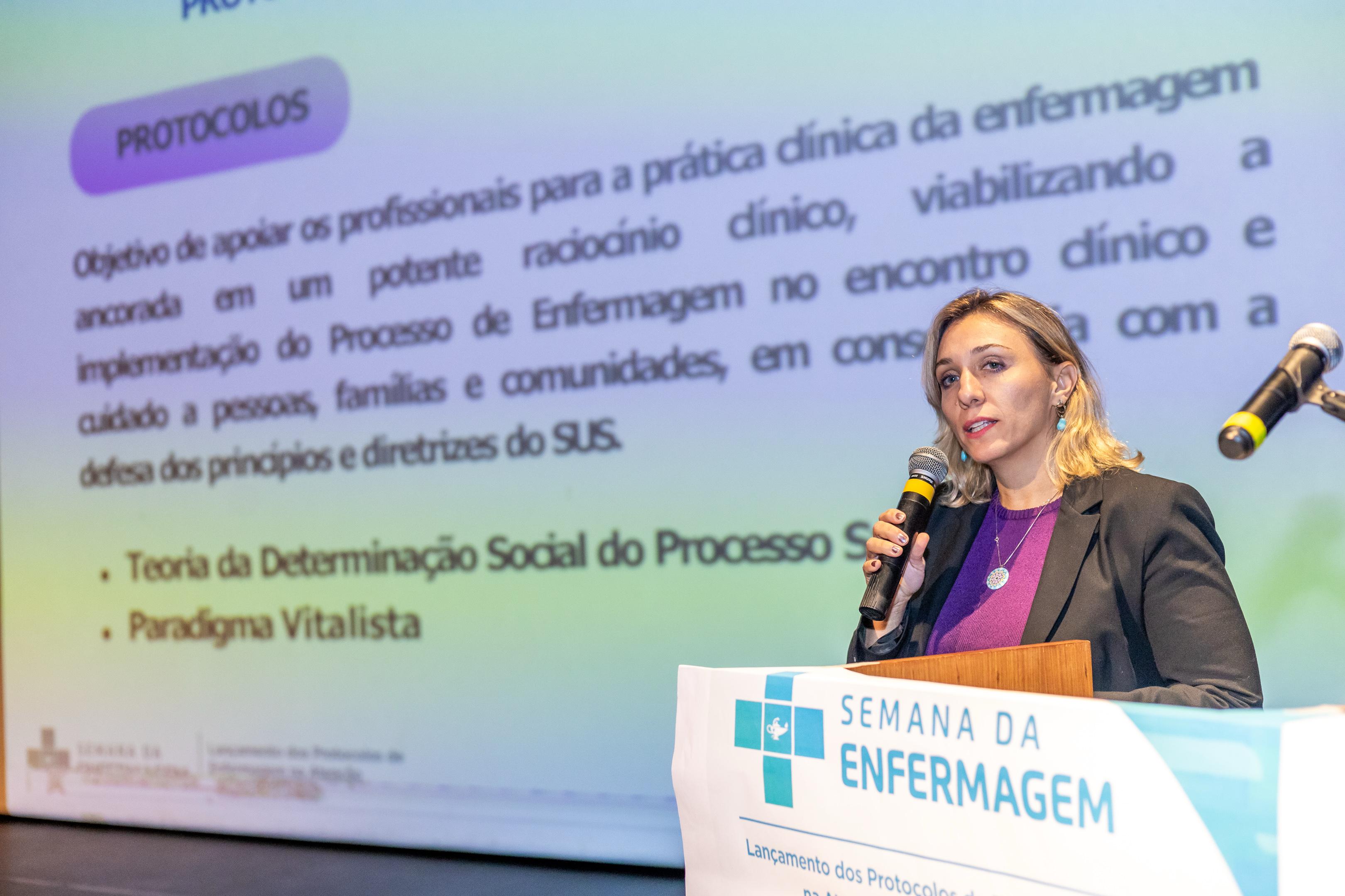 A imagem mostra uma mulher palestrando durante um evento da Semana da Enfermagem, identificado por uma faixa à frente do púlpito onde se lê: