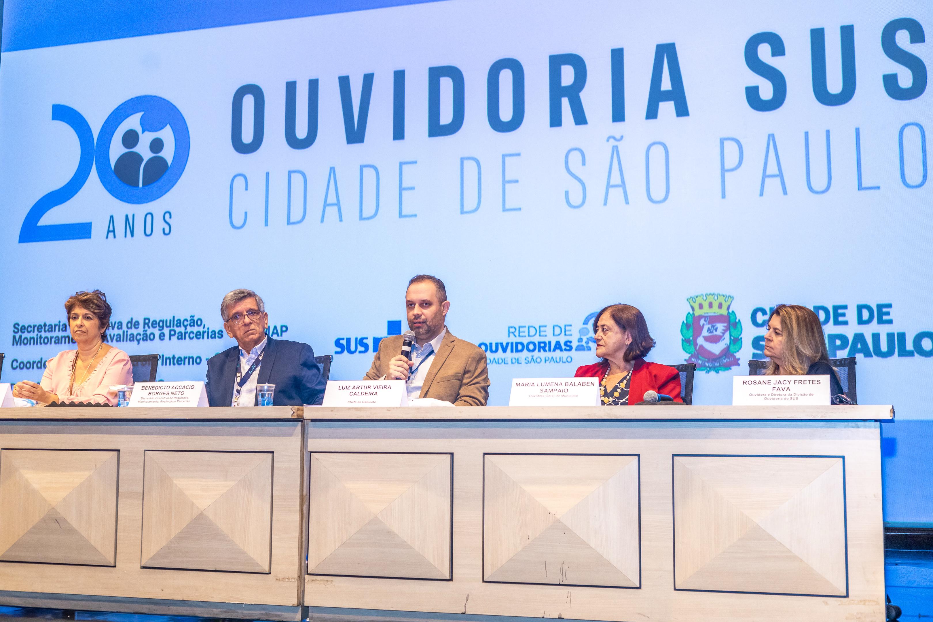 A imagem mostra uma mesa de conferência com cinco pessoas sentadas, participando de um evento em comemoração aos 20 anos da Ouvidoria SUS da Cidade de São Paulo. Ao fundo, há um painel com a identidade visual do evento e logotipos da Cidade de São Paulo, SUS, Secretaria Municipal da Saúde (SMS-SP), entre outros.  Da esquerda para a direita, os participantes são:  Uma mulher de roupa clara;  Benedicto Accacio Borges Neto (identificado pela placa), provavelmente representando um órgão de controle interno;  Luiz Artur Vieira Caldeira, que segura um microfone e parece estar falando – identificado como chefe de gabinete da Secretaria Municipal da Saúde;  Maria Lumena Balaben Sampaio, também identificada por uma placa;  Rosane Jacy Fretes Fava, da Ouvidoria-Geral do SUS (Ministério da Saúde).  O evento celebra e reforça a importância da ouvidoria como canal de escuta e participação cidadã no SUS paulistano.