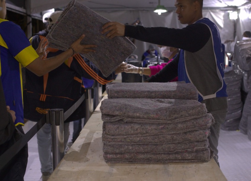 A imagem mostra uma cena de distribuição de cobertores em um ambiente que aparenta ser uma estrutura montada para atendimento emergencial ou ação social, como uma tenda. No primeiro plano, vemos uma pilha de cobertores dobrados sobre uma mesa. À direita, um homem usando colete azul entrega um cobertor a uma pessoa do outro lado da mesa. Outras pessoas com coletes e equipamentos de proteção, como máscaras, também estão envolvidas na ação