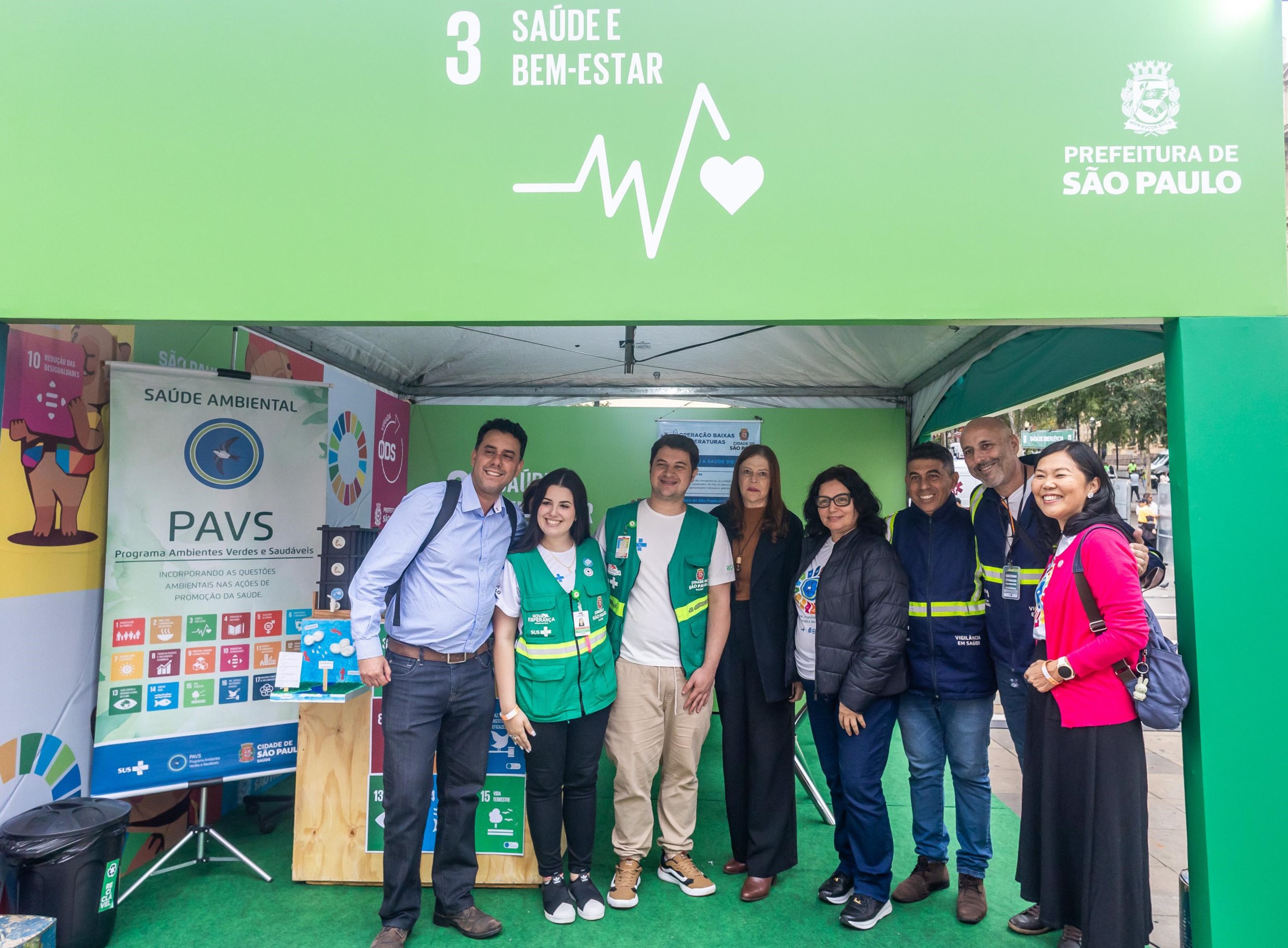A imagem mostra um grupo de nove pessoas posando sorridentes em frente a uma tenda verde identificada com o símbolo do Objetivo de Desenvolvimento Sustentável (ODS) 3 – Saúde e Bem-Estar, da Prefeitura de São Paulo. A tenda abriga o estande do Programa Ambientes Verdes e Saudáveis (PAVS), ligado à Saúde Ambiental, como indica o banner exposto no local.  Os integrantes do grupo estão vestidos com diferentes trajes: alguns com coletes verdes do programa, outros com uniformes azuis da vigilância sanitária e trajes casuais. A ambientação inclui cartazes coloridos com os ícones dos ODS, destacando metas como saúde, educação, igualdade de gênero, cidades sustentáveis, entre outros. O cenário urbano e a arquitetura ao fundo sugerem que o evento ocorre em um espaço público da cidade.