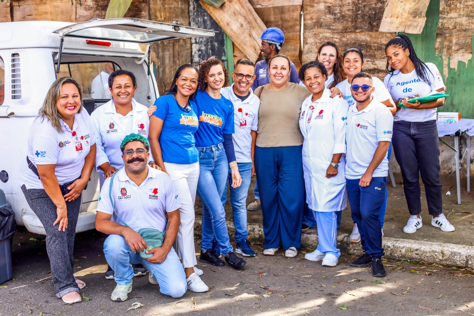 A imagem mostra um grupo de profissionais do Consultório na Rua e da Uvis Capela do Socorro reunidos ao ar livre, posando para a foto com expressões alegres. Ao todo, há treze pessoas visíveis em destaque, com diferentes uniformes e camisetas identificando a atuação na área da saúdePrimeiro plano, à esquerda: Uma mulher de pele clara, com cabelo loiro e liso na altura dos ombros, está sorrindo. Ela usa camiseta branca com logotipos do SUS e do programa “Imuniza Capela”. Ao lado dela, uma mulher de pele parda com cabelo preso e jaleco branco também sorri para a foto.Ao centro, na frente do grupo, agachado: Um homem de pele parda, com cabelo verde curto e cacheado e um grande bigode preto, usa camiseta branca do SUS e segura um boné verde. Ele também sorri.No centro, em pé: Duas mulheres com camisetas azuis com a inscrição “Imuniza Capela” estão lado a lado, sorrindo. Ambas têm cabelos cacheados, uma com cabelo curto e a outra médio. À direita delas, uma mulher de pele parda com cabelo escuro preso usa blusa bege e calça azul larga. À direita do grupo: Uma mulher negra de cabelo preso usa jaleco branco e sorri amplamente.Um homem de baixa estatura, pele clara e óculos escuros, com camiseta do SUS e calça azul, sorri ao lado dela. Três mulheres ao fundo completam o grupo, duas delas com camisetas brancas, e uma com prancheta verde e calça jeans escura.O grupo está em frente a uma Kombi branca com a porta traseira aberta. Ao fundo, há um muro de madeira e concreto. O ambiente é externo