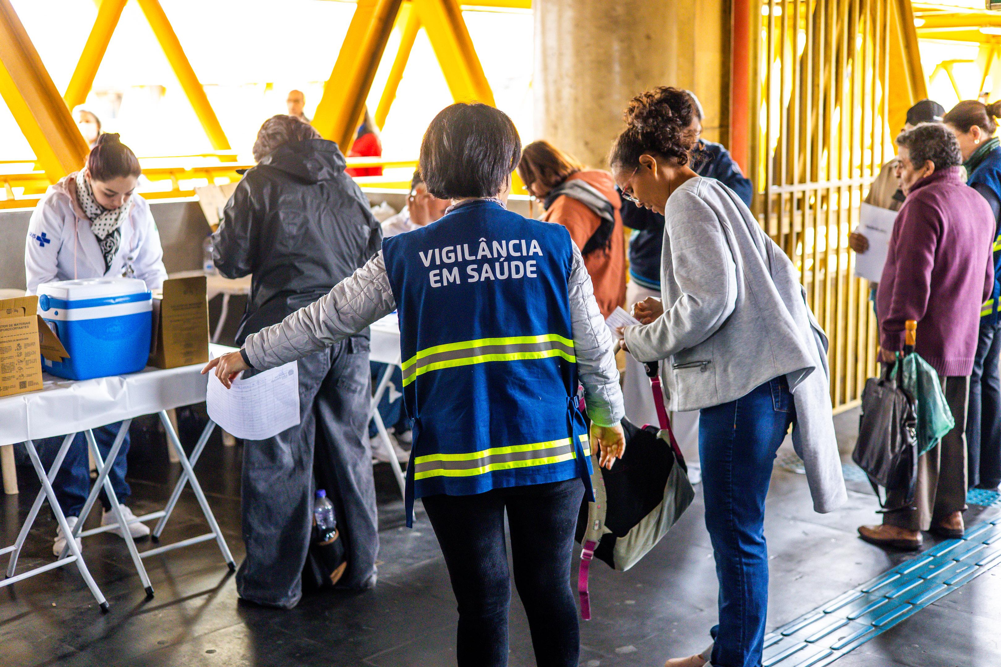 A imagem mostra uma ação de saúde pública em um espaço coberto, possivelmente uma estação ou terminal de transporte. Em primeiro plano, uma agente de saúde veste um colete azul com a inscrição “VIGILÂNCIA EM SAÚDE” e está orientando uma mulher que segura uma bolsa e um papel. À esquerda, profissionais de saúde trabalham em mesas dobráveis cobertas com toalhas brancas, onde há caixas de papelão e uma caixa térmica azul, provavelmente para armazenamento de vacinas ou medicamentos. Ao fundo, várias pessoas aguardam atendimento em fila. O ambiente é bem iluminado e a estrutura metálica amarela indica um local urbano de grande circulação. A cena transmite organização e cuidado com a saúde coletiva.