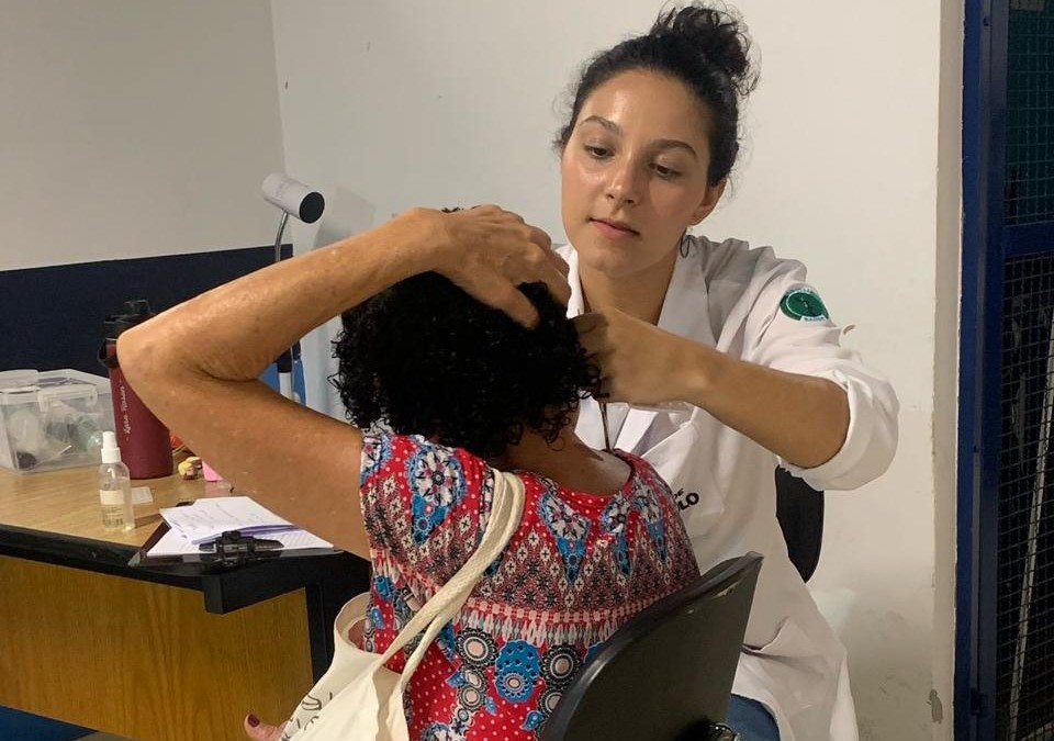 A imagem mostra uma profissional da área da saúde realizando atendimento a uma mulher. A profissional está usando um jaleco branco e parece estar examinando a orelha ou a cabeça da paciente, que está sentada de costas, com as mãos levantadas para cima da cabeça, facilitando o acesso da examinadora. A paciente veste uma blusa estampada com padrões coloridos e carrega uma bolsa de tecido no ombro. O ambiente parece ser uma sala de atendimento simples, com uma mesa ao fundo contendo alguns materiais, como papéis, garrafa térmica, frascos e caixas organizadoras. A parede ao fundo é branca e azul escuro, e há um refletor ou abajur preso à mesa. A cena transmite um momento de cuidado e atenção em um contexto de saúde