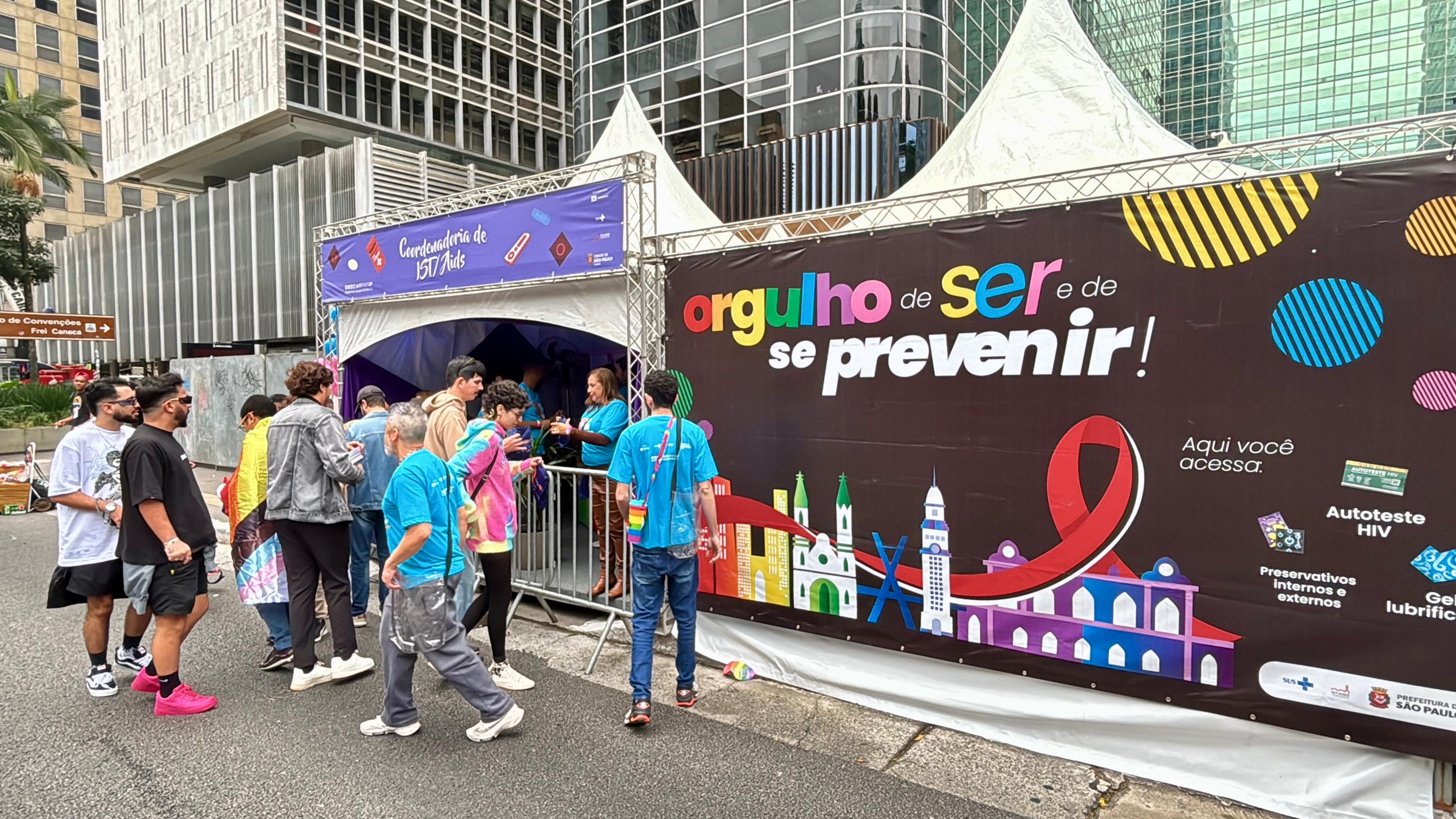 A imagem mostra uma tenda de atendimento da Coordenadoria de ISTs/Aids da cidade de São Paulo montada em via pública, durante a Parada do Orgulho LGBT+. A tenda branca tem um letreiro superior roxo com ícones de preservativos, comprimidos e seringas, e está cercada por grades metálicas para organização da fila. Na lateral da tenda, há um grande painel colorido com os dizeres: