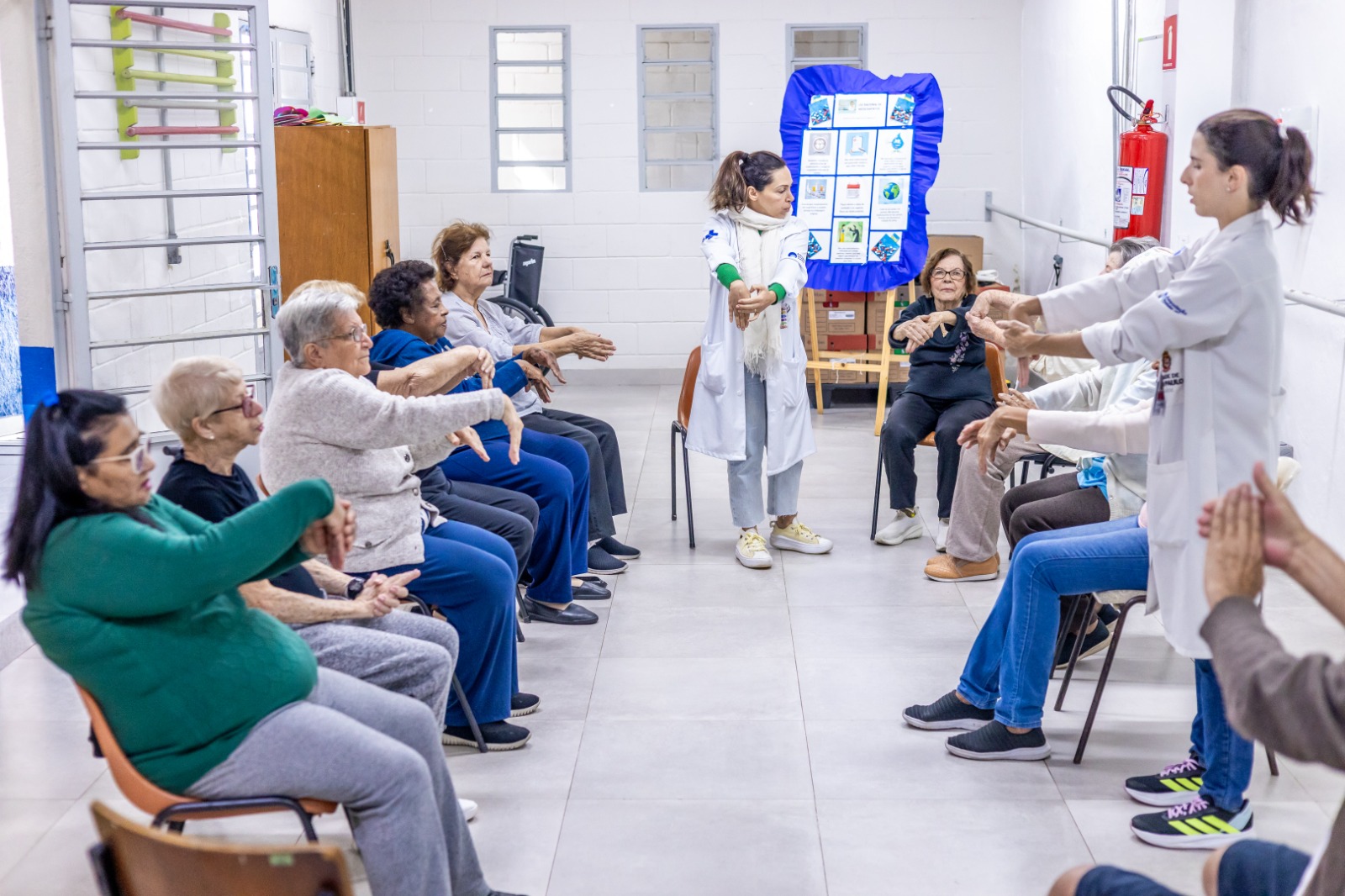 A imagem mostra uma sala de atividades coletivas onde um grupo de idosos participa de uma sessão de alongamento. Os participantes, sentados em cadeiras dispostas em semicírculo, estão com os braços estendidos à frente, fazendo movimentos com os punhos e mãos, sob a orientação de duas profissionais de saúde. As profissionais — ambas vestindo jalecos brancos — estão de pé, demonstrando os exercícios. Uma delas usa calça clara, tênis brancos, camiseta verde e um cachecol branco; a outra está do lado direito da imagem, com o cabelo preso e calça jeans azul. O ambiente é bem iluminado, com paredes brancas, piso claro e janelas pequenas ao fundo. Há um painel informativo coberto com tecido azul na parte de trás da sala, além de um armário e um extintor de incêndio visíveis nas laterais. Os idosos presentes são homens e mulheres, de diferentes tons de pele, com cabelos grisalhos ou brancos, vestindo roupas confortáveis. A atmosfera é acolhedora e de cuidado, sugerindo um espaço voltado à promoção da saúde e bem-estar na terceira idade.