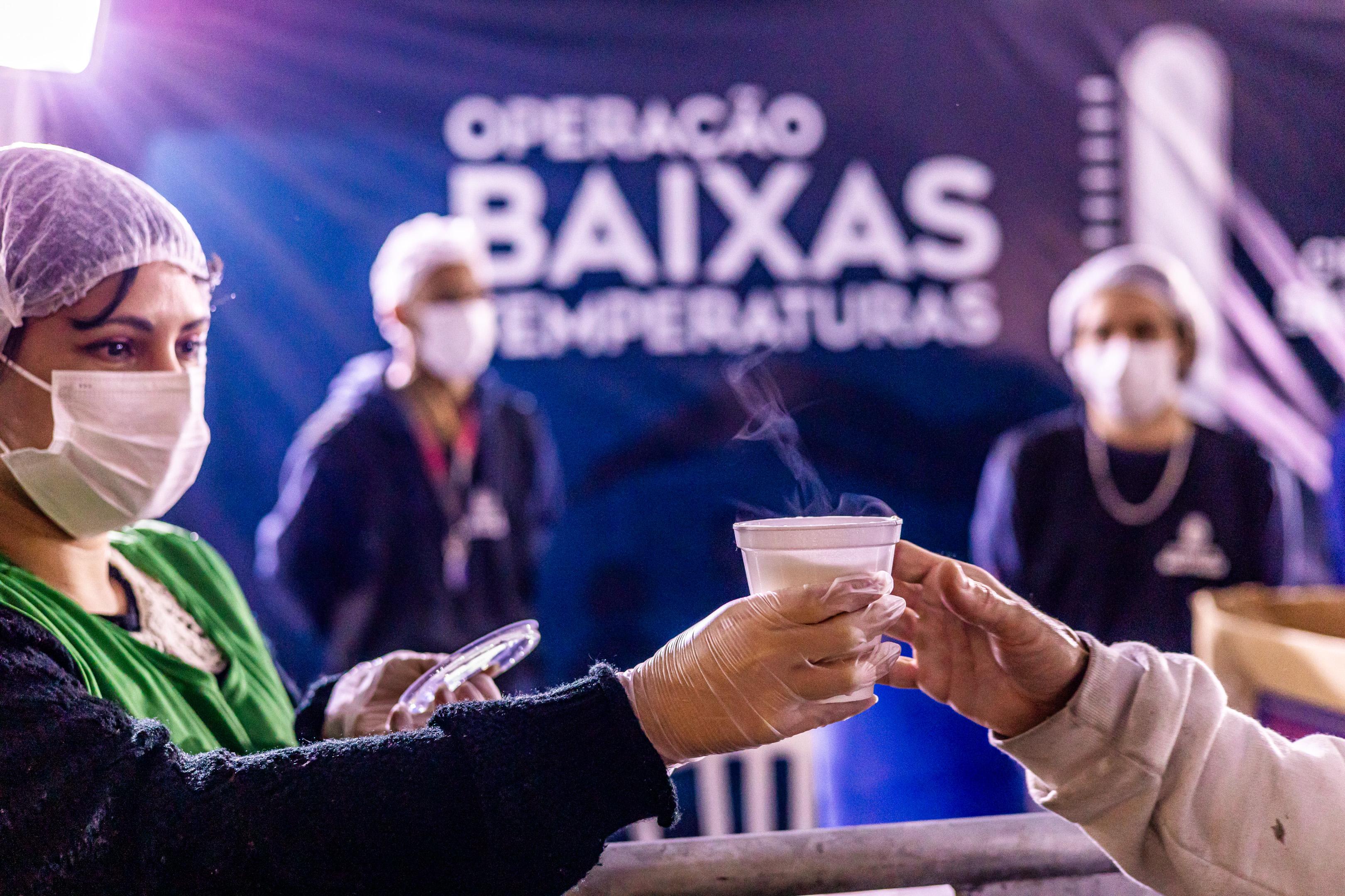A imagem mostra uma cena de acolhimento durante a Operação Baixas Temperaturas, identificada por uma faixa no fundo. Em primeiro plano, uma profissional de saúde ou assistência social — usando touca, máscara e luvas — entrega um copo de bebida quente a uma pessoa. O vapor visível indica que o líquido está recém-preparado, sugerindo cuidado com o conforto térmico dos atendidos. Ao fundo, outras pessoas com uniforme e máscara reforçam a ideia de uma equipe organizada e mobilizada para apoiar populações vulneráveis em dias de frio intenso. A atmosfera transmite solidariedade, acolhimento e proteção.